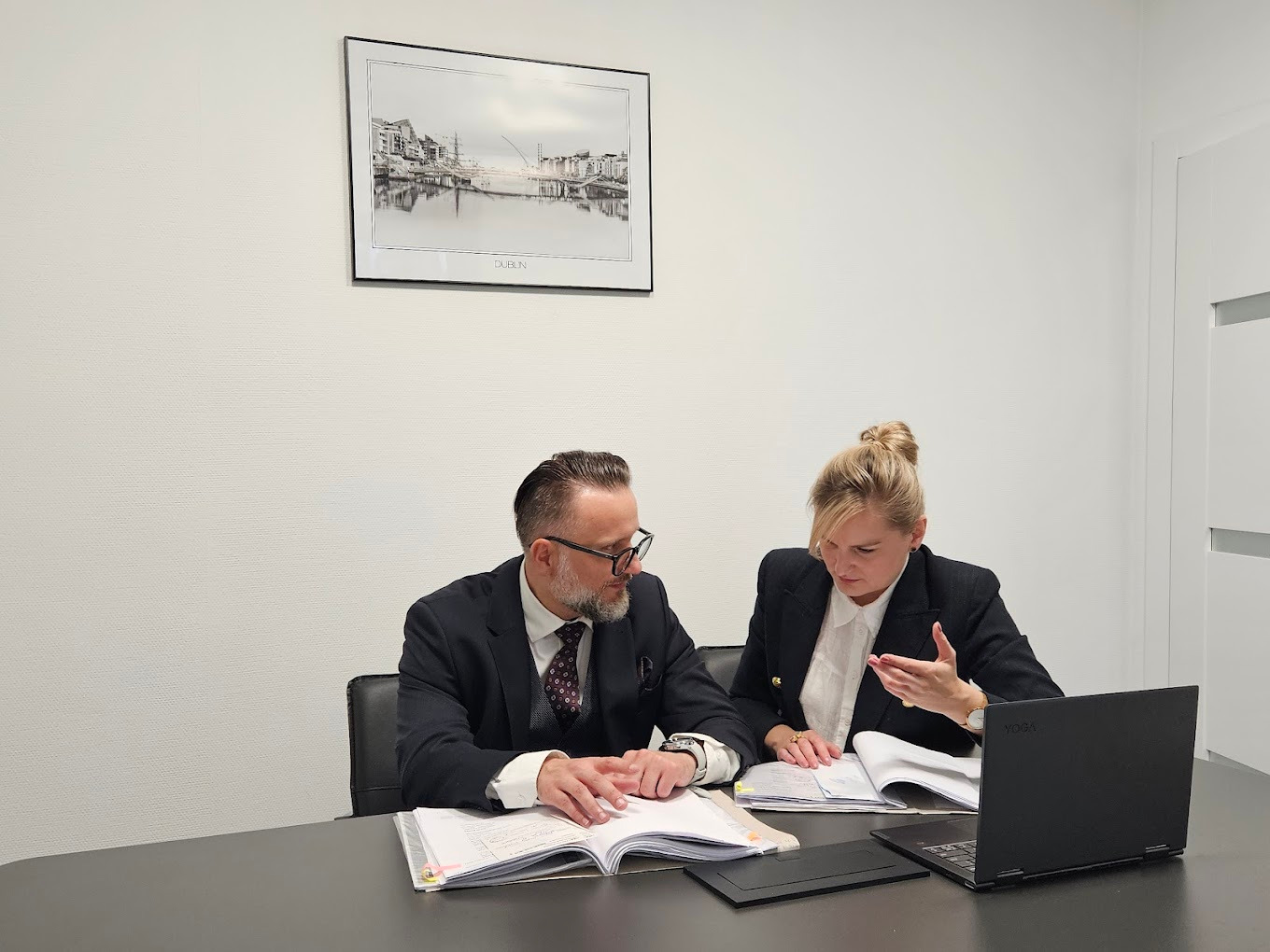 Dwoje prawników w biurze analizuje dokumenty przy biurku, na którym leży laptop. Na ścianie wisi obraz z motywem Dublina.