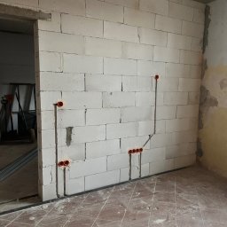 VRWOLT - Usługi Elektryczne - Ściana z betonu komórkowego z wyżłobieniami pod przewody i puszkami elektrycznymi. Widoczne bruzdy pionowe i poziome. W tle fragment pomieszczenia w trakcie remontu.