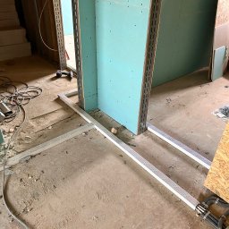VRWOLT - Usługi Elektryczne - Montaż ścianki działowej z płyt gipsowo-kartonowych na metalowym stelażu. Widoczne kable w peszlach. W tle fragment schodów i narzędzia.