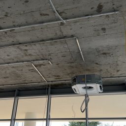 VRWOLT - Usługi Elektryczne - Montaż instalacji elektrycznej pod sufitem w budynku komercyjnym. Widoczne rury i kable, klimatyzator. Prace instalacyjne w trakcie realizacji.