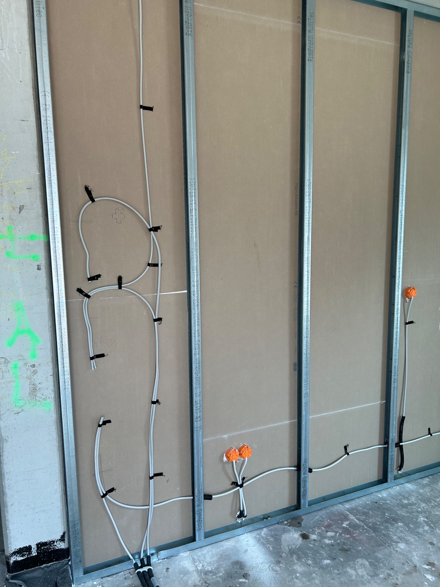 Białe przewody elektryczne przytwierdzone czarnymi opaskami do metalowego stelażu na tle płyty kartonowo-gipsowej, widoczne pomarańczowe puszki instalacyjne.