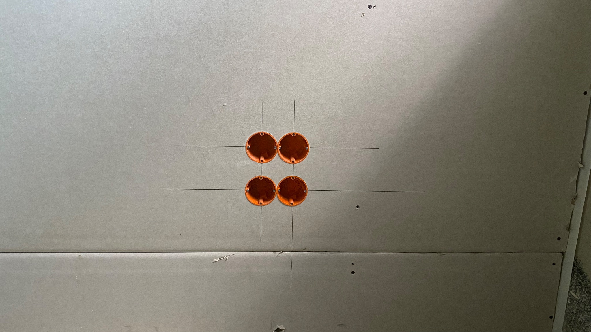 Cztery okrągłe, pomarańczowe puszki elektryczne zamontowane w szarej płycie gipsowo-kartonowej, z widocznymi liniami pomocniczymi wyznaczającymi ich położenie.