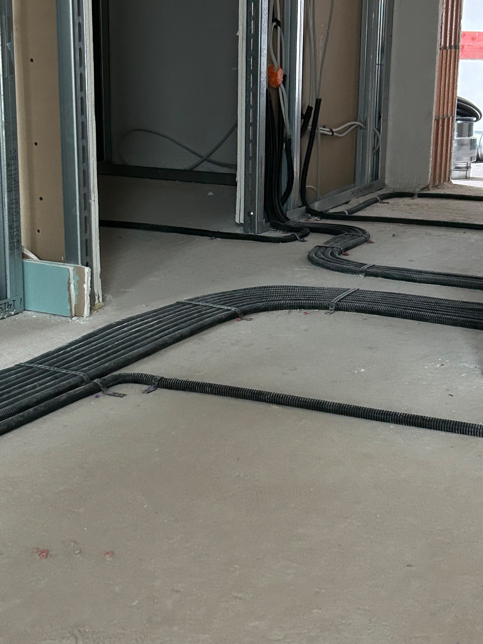 Rury instalacji elektrycznej ułożone równolegle na surowym betonie w trakcie prac wykończeniowych, widoczne profile konstrukcyjne ścian.