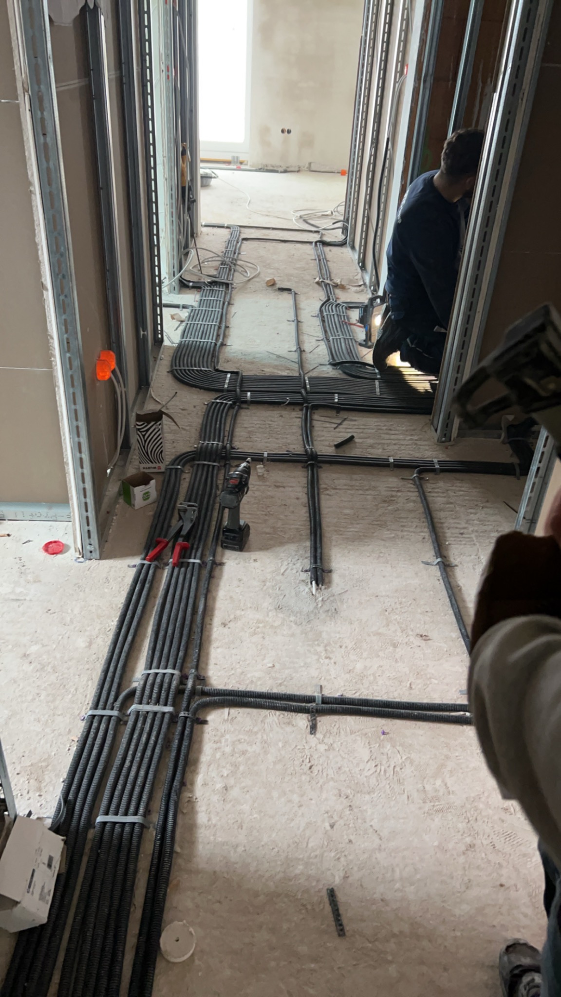 Korytarz w trakcie remontu z widocznymi przewodami elektrycznymi ułożonymi na podłodze, zabezpieczonymi opaskami zaciskowymi, w tle instalator w trakcie pracy.