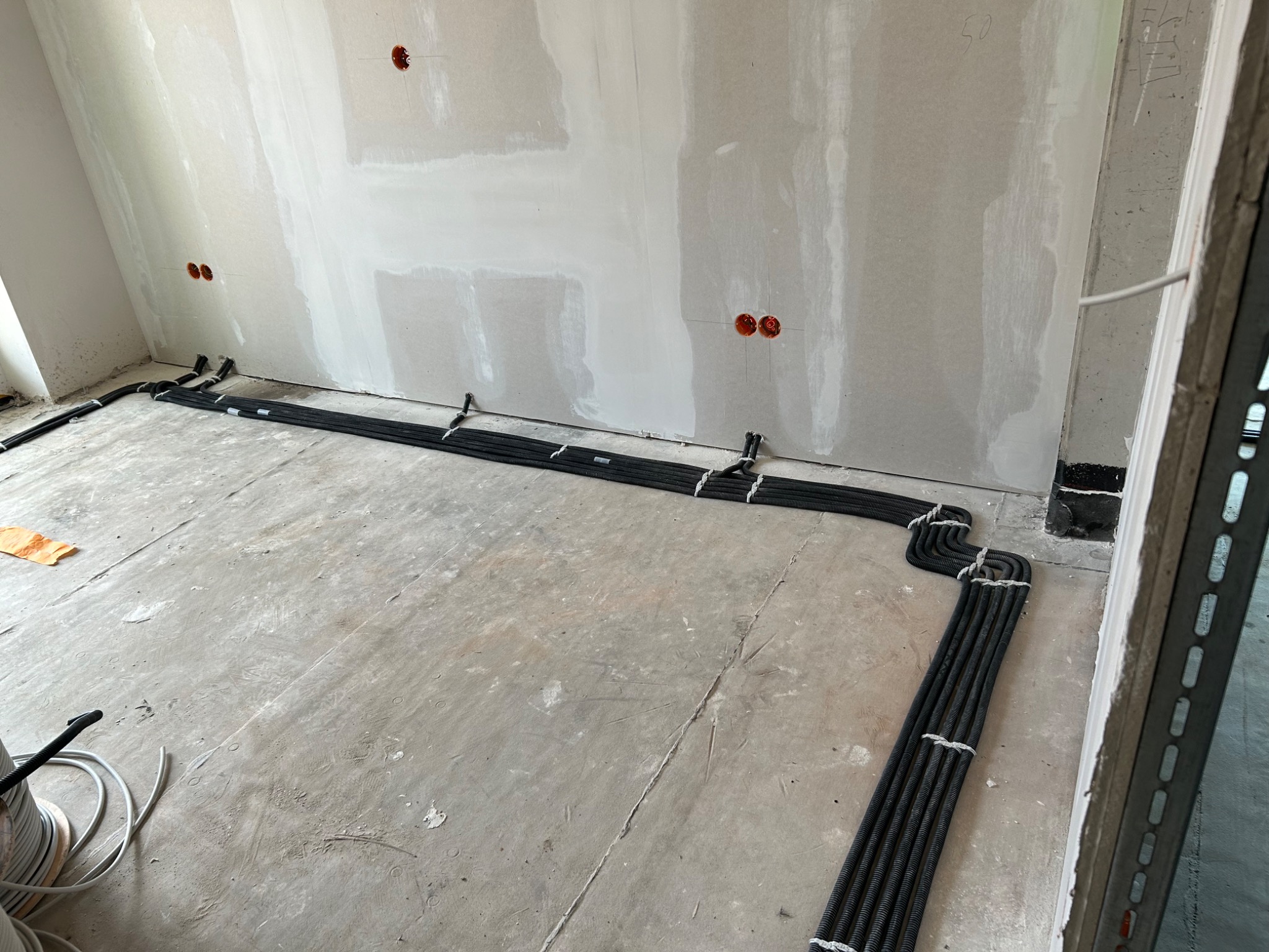 Świeżo położone, czarne kable elektryczne w peszlach, ułożone równolegle na surowej podłodze w nowo wykończonym pomieszczeniu z płyt kartonowo-gipsowych, widoczne puszki instalacyjne w ścianie.