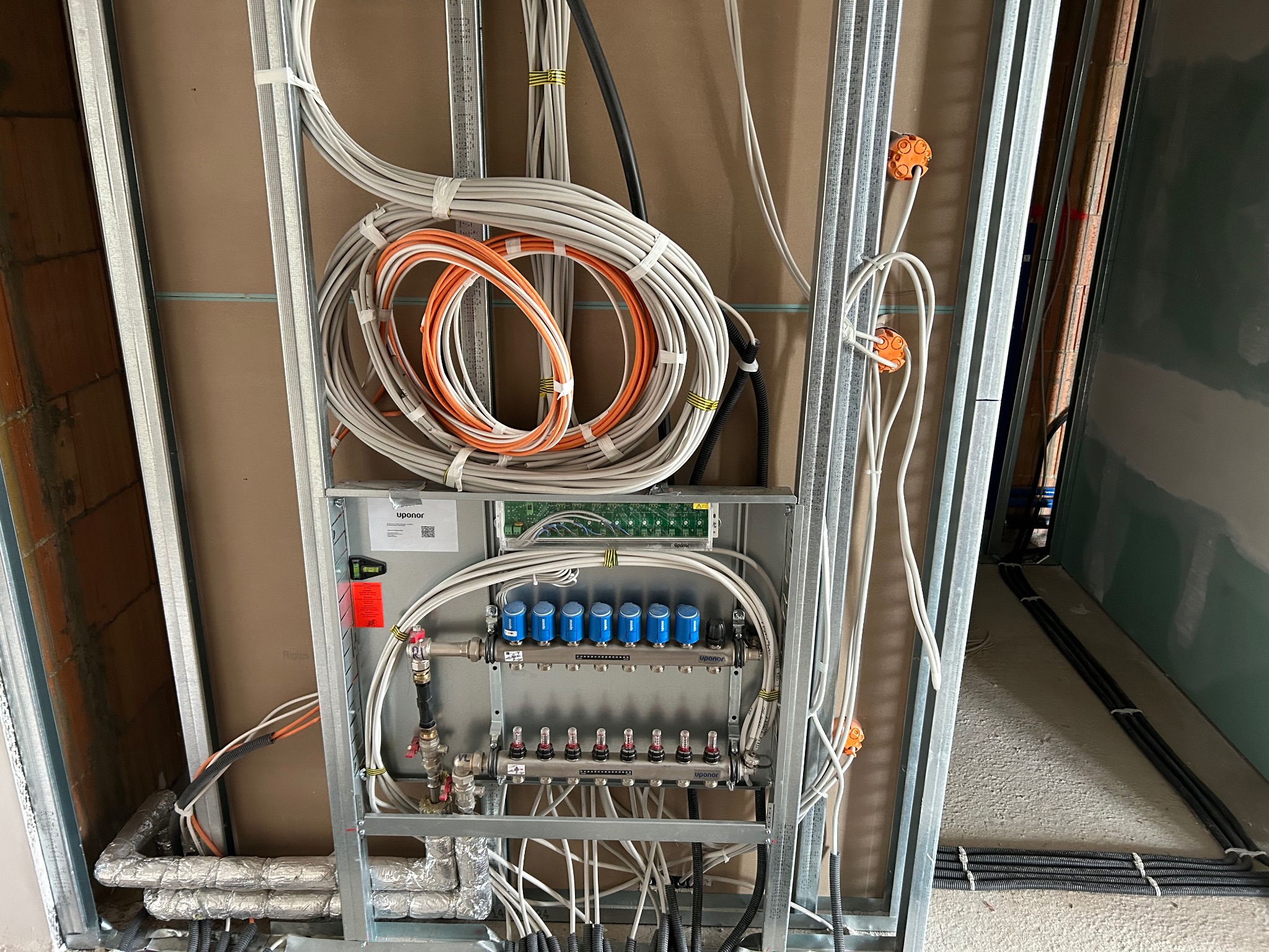 Rozdzielacz ogrzewania podłogowego Uponor w trakcie montażu, widoczne liczne przewody elektryczne i rury grzewcze w metalowej konstrukcji szkieletowej ściany.