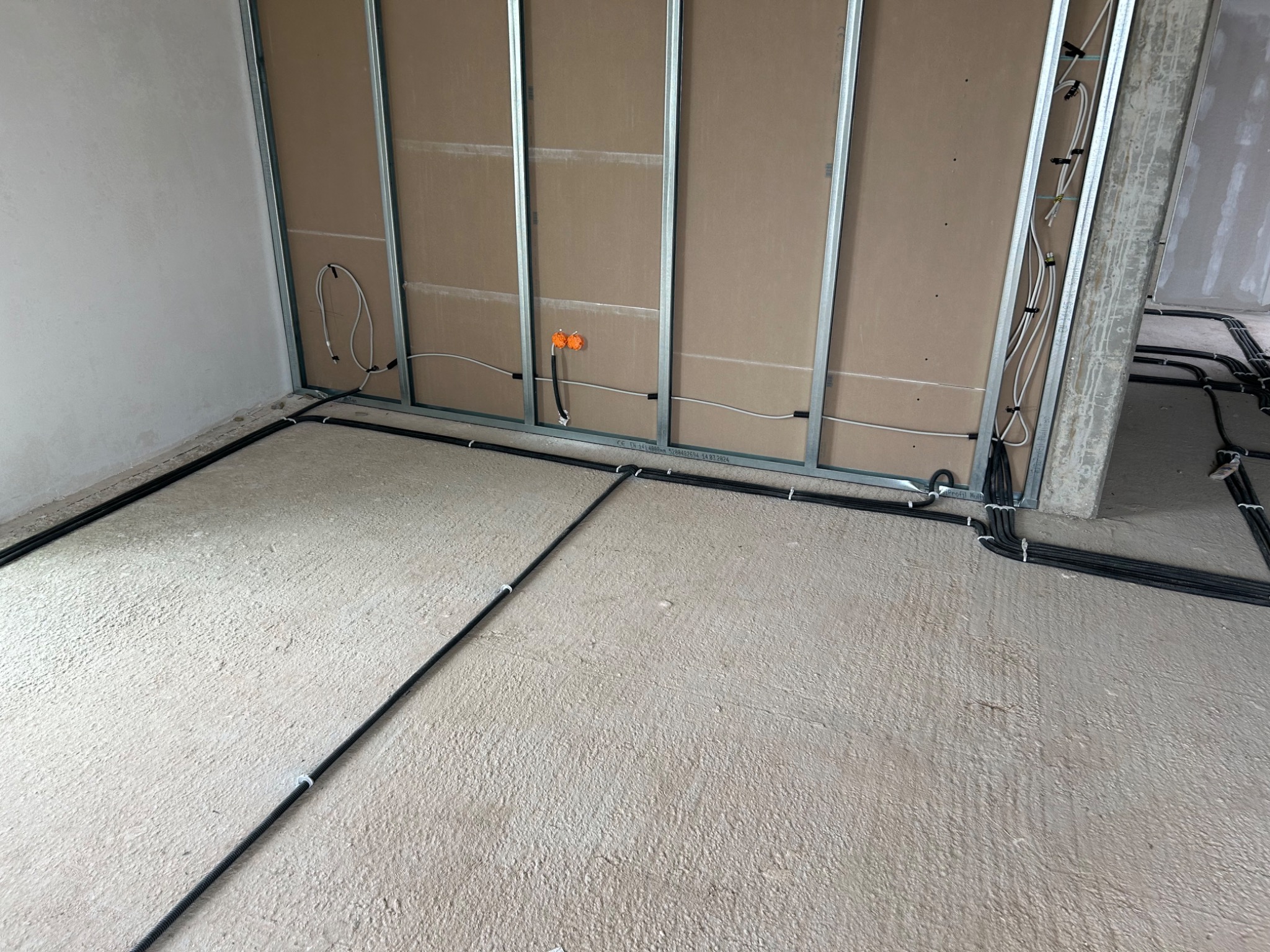 Surowe wnętrze w trakcie prac wykończeniowych; widoczne kable elektryczne poprowadzone po podłodze i wzdłuż metalowej konstrukcji ścian działowych z płyt kartonowo-gipsowych; pomarańczowe puszki...