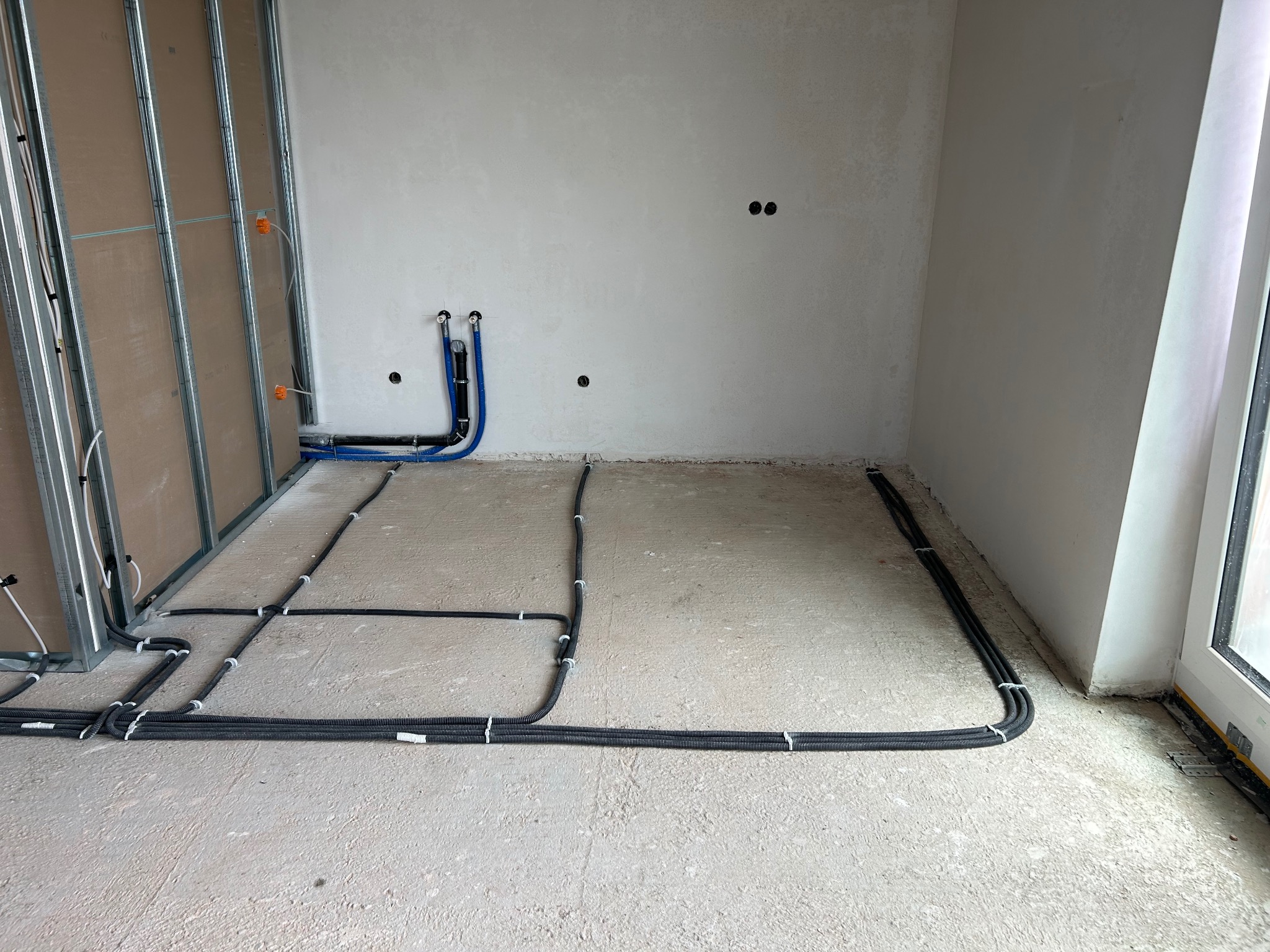 Surowe wnętrze w trakcie prac wykończeniowych: widoczne kable elektryczne w peszlach ułożone na podłodze, rury doprowadzające wodę do ściany, metalowa konstrukcja pod zabudowę z płyt...