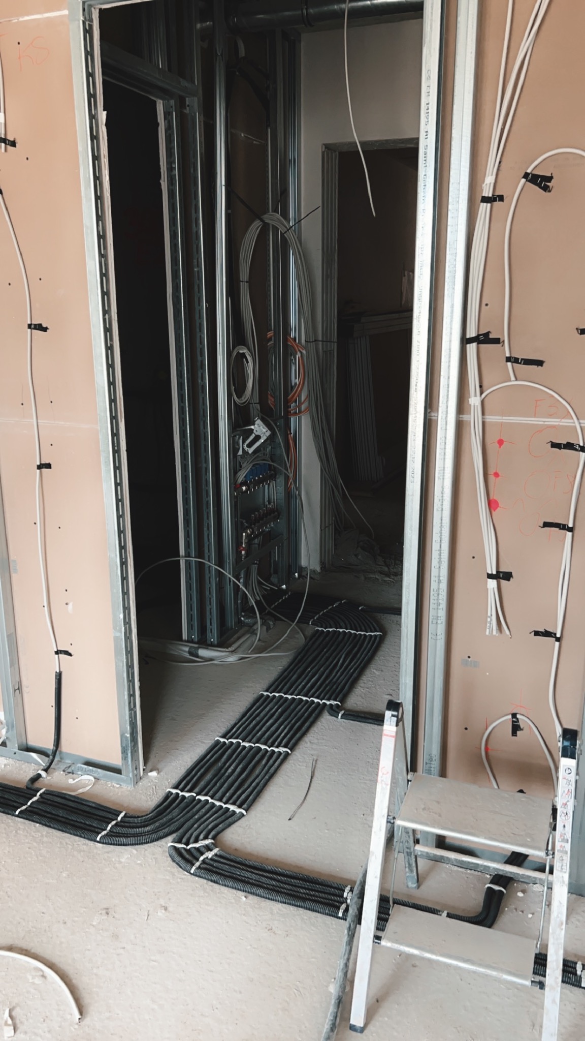 Wiązka czarnych przewodów w peszlu ułożona na podłodze w surowym stanie, widoczna drabina aluminiowa i instalacja elektryczna w ścianie z profili, w tle otwór drzwiowy.