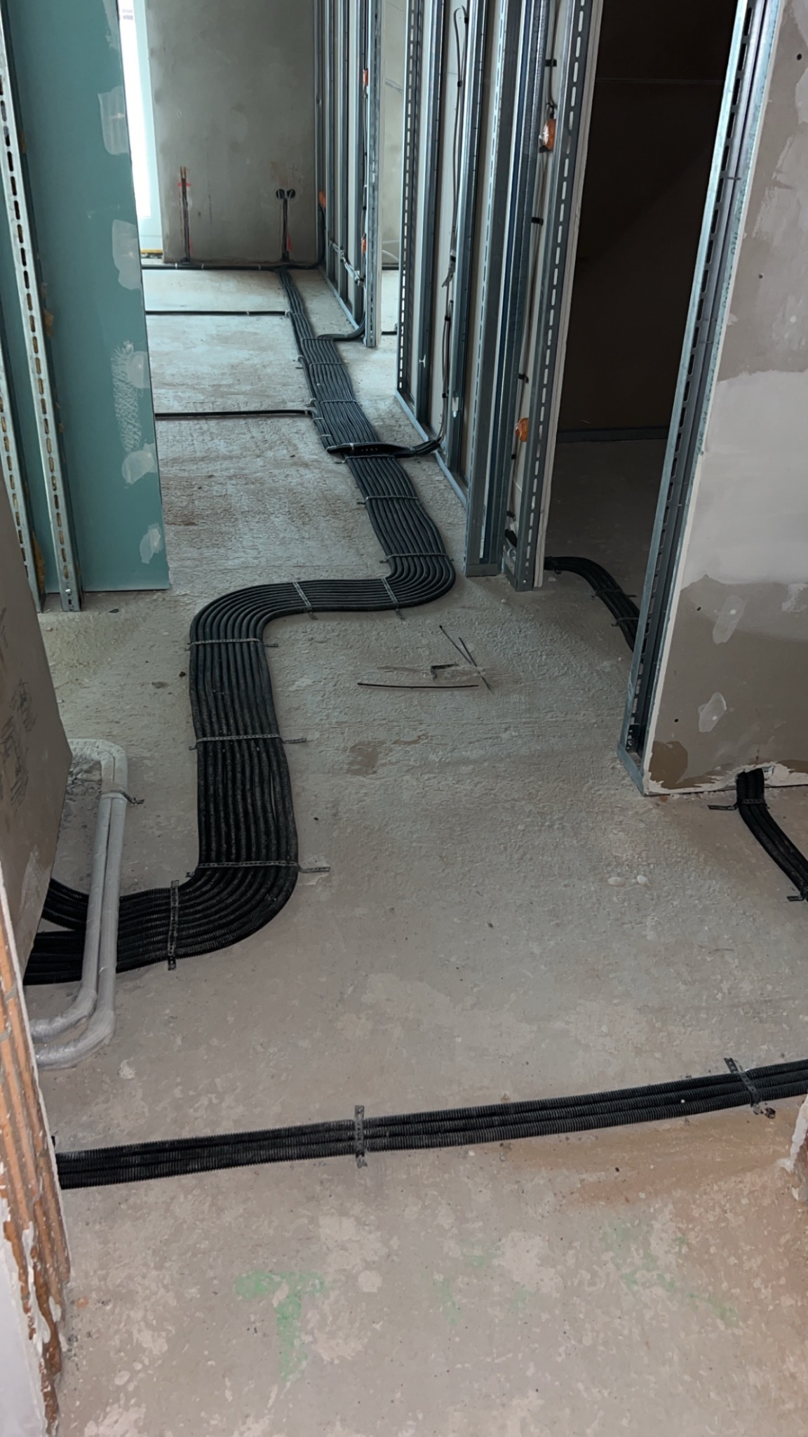 Rury ochronne kabli elektrycznych ułożone na podłodze w surowym stanie, prowadzone wzdłuż metalowych profili ścian działowych w trakcie budowy.