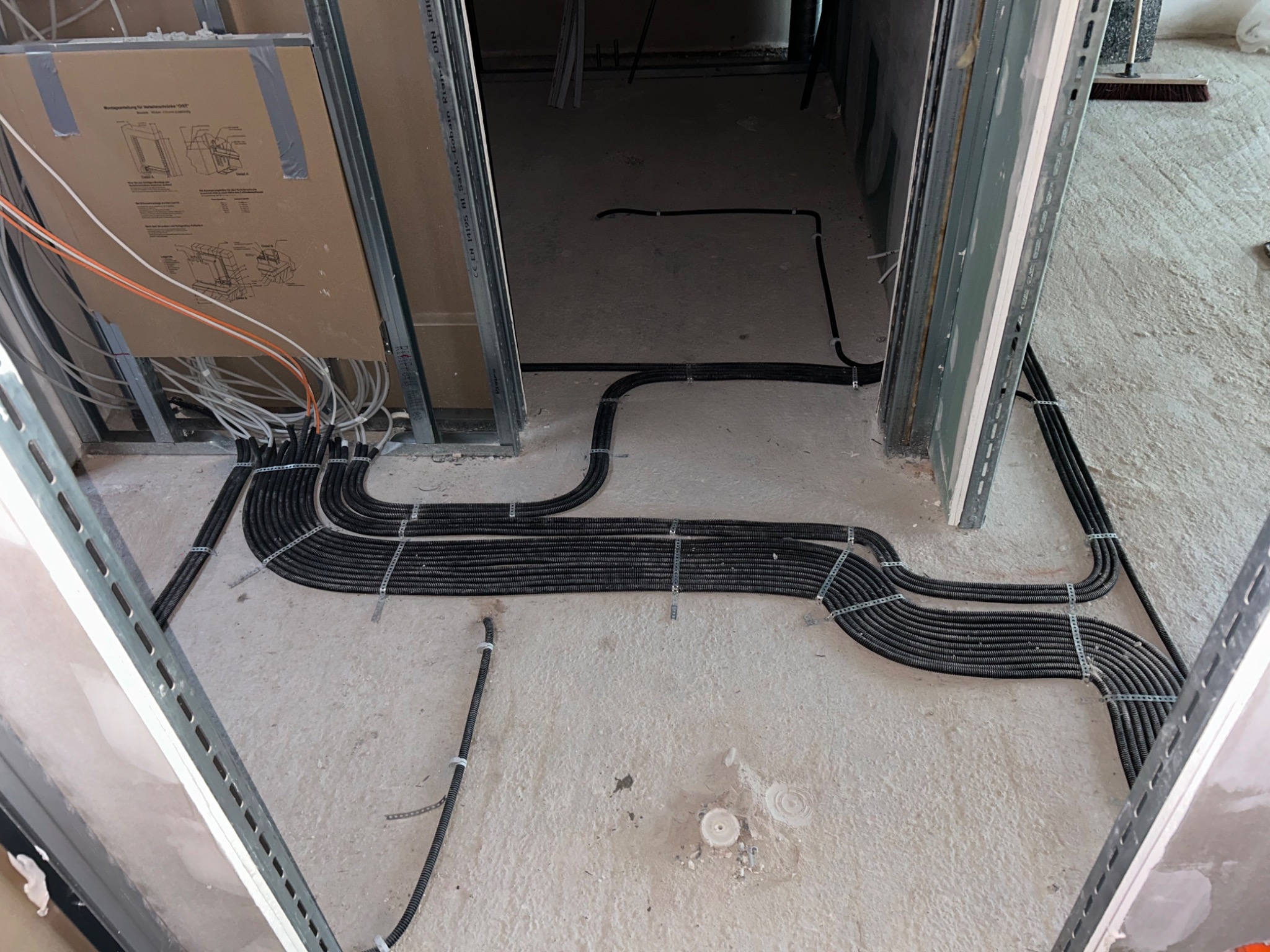 Równolegle ułożone, czarne rury karbowane z przewodami elektrycznymi, mocowane metalowymi obejmami do surowej, betonowej podłogi w trakcie prac instalacyjnych wewnątrz budynku.