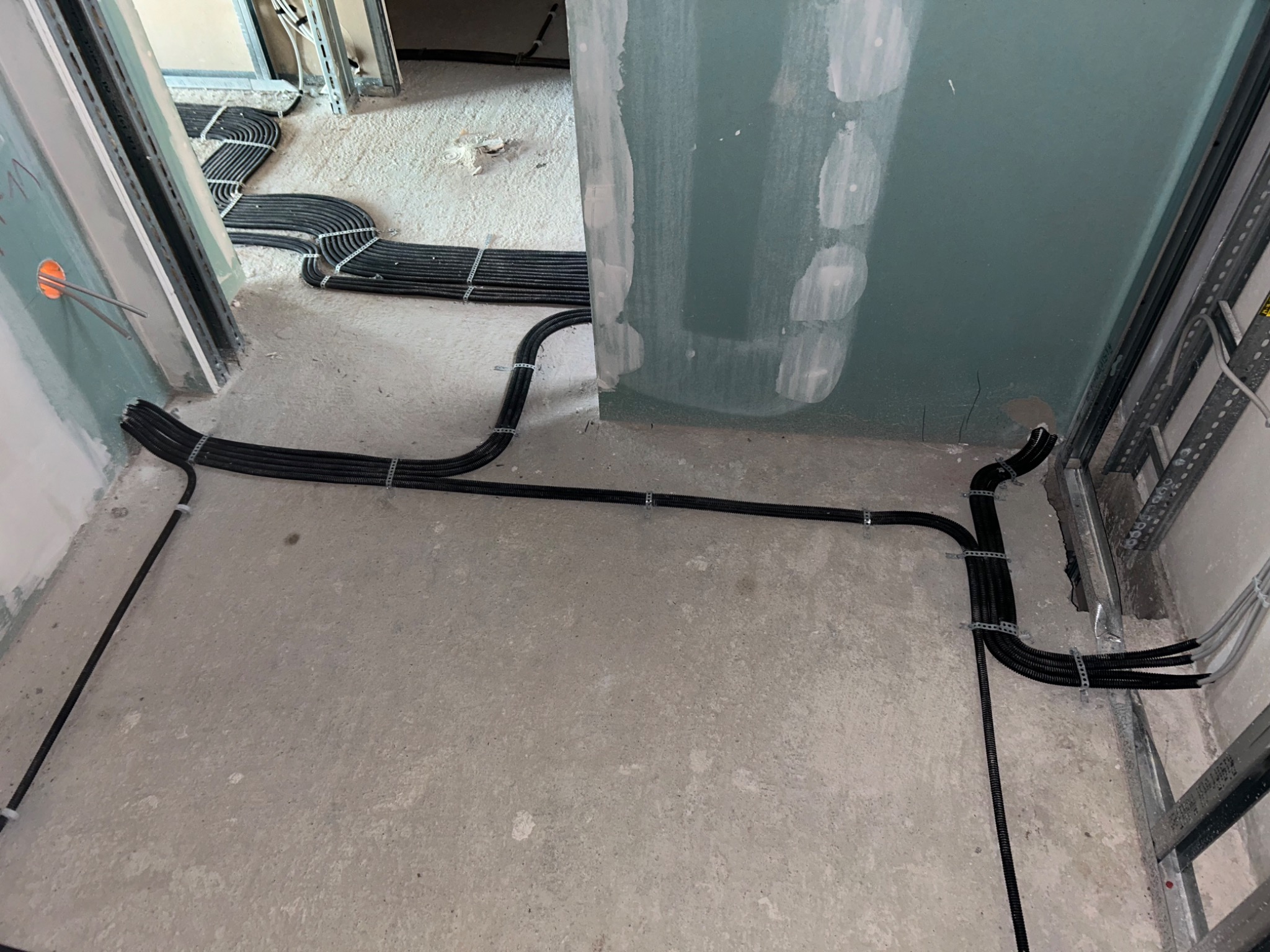 Czarny peszel z kablami elektrycznymi ułożony na surowym betonowym podłożu w trakcie prac instalacyjnych, przymocowany metalowymi uchwytami do podłoża i ściany z płyt gipsowo-kartonowych.