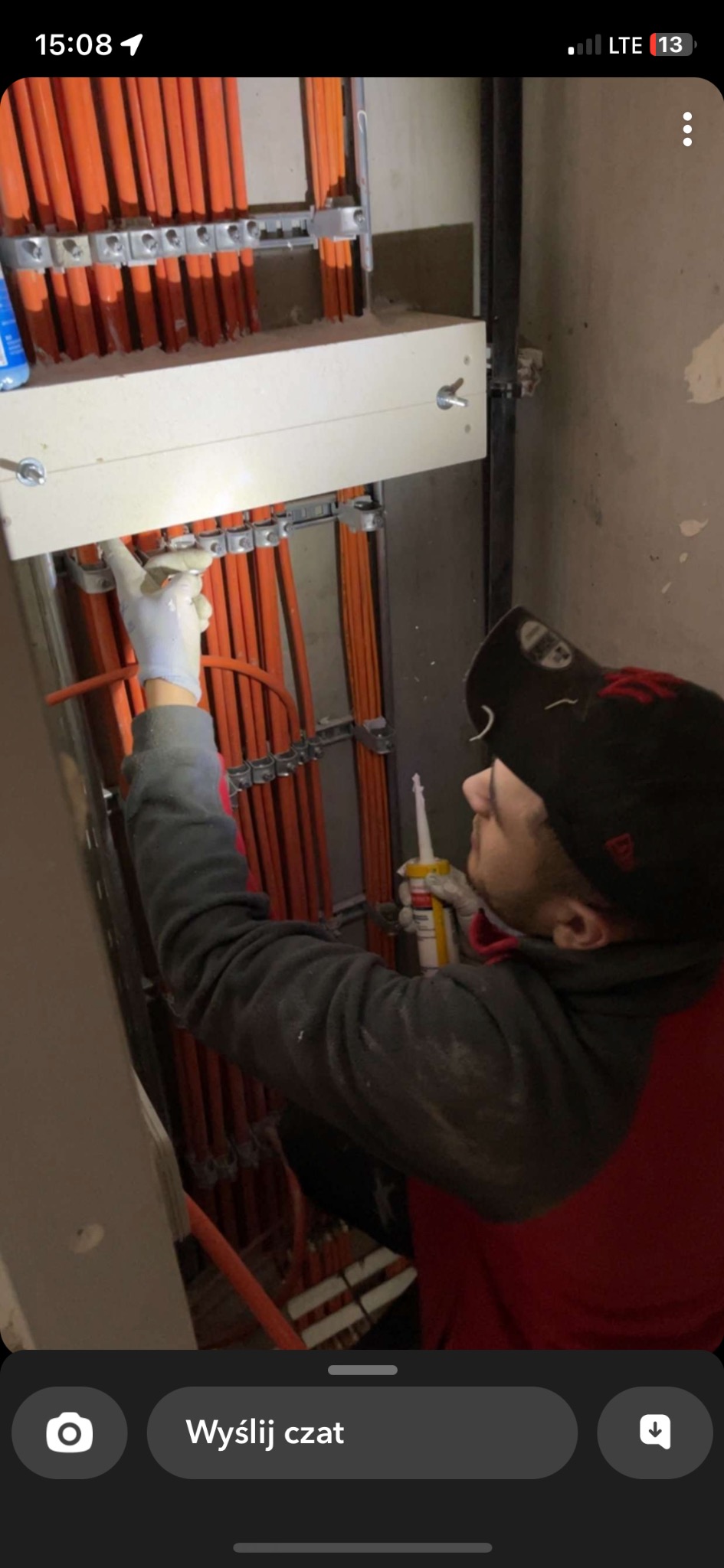 Elektryk w rękawicach uszczelnia silikonem pomarańczowe przewody elektryczne zamocowane na metalowych uchwytach do ściany.