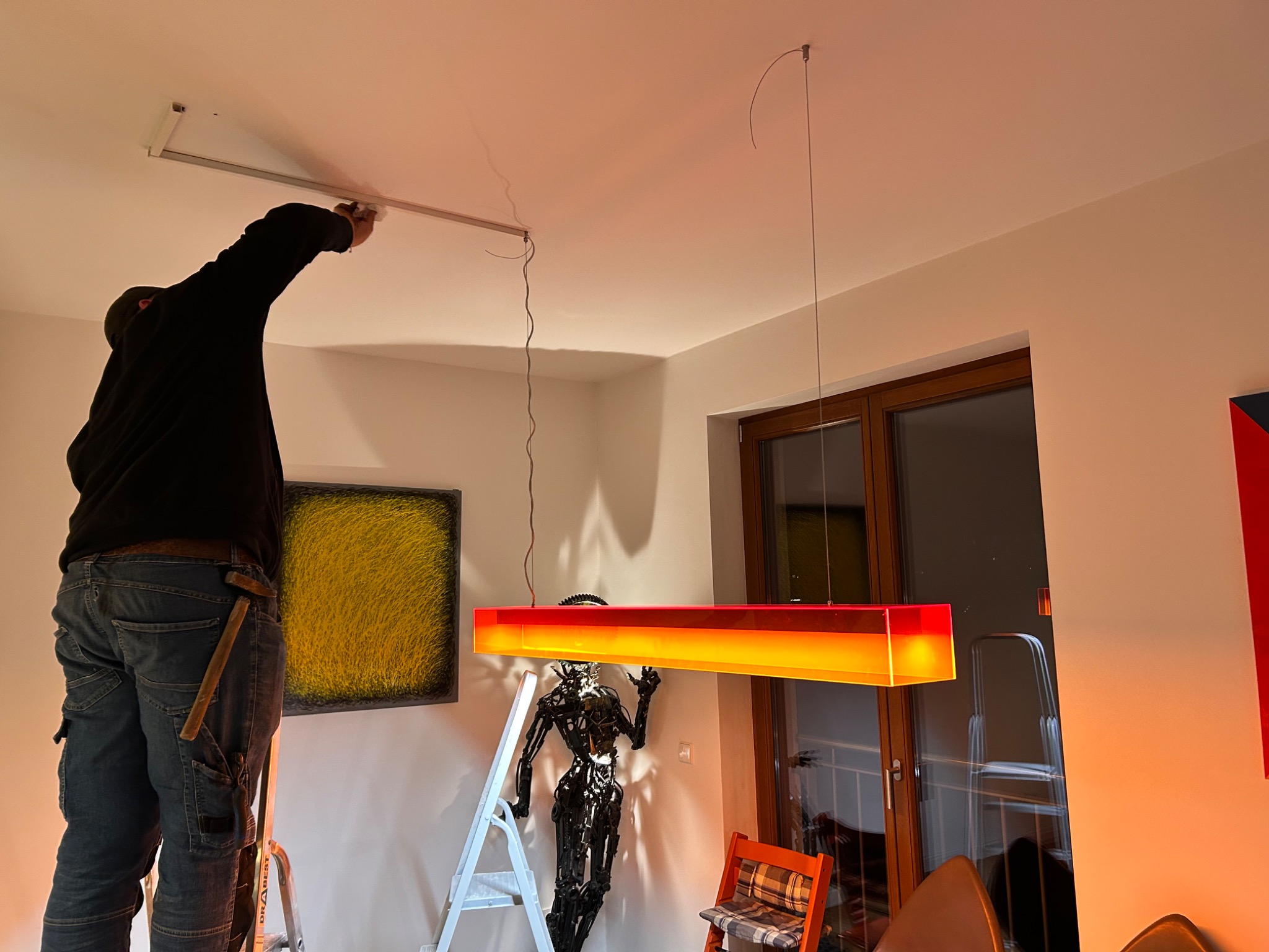 Elektryk na drabinie montuje oświetlenie sufitowe w nowoczesnym wnętrzu. Widoczna lampa wisząca i dekoracje. Nietypowe ujęcie z narzędziem w kieszeni.