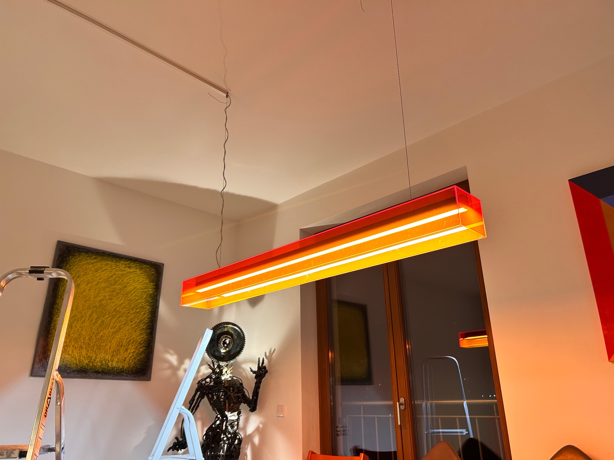 Pomarańczowa lampa LED w nowoczesnym wnętrzu, widoczna drabina i dekoracyjna rzeźba. Oświetlenie sufitowe z elementami czerwieni.