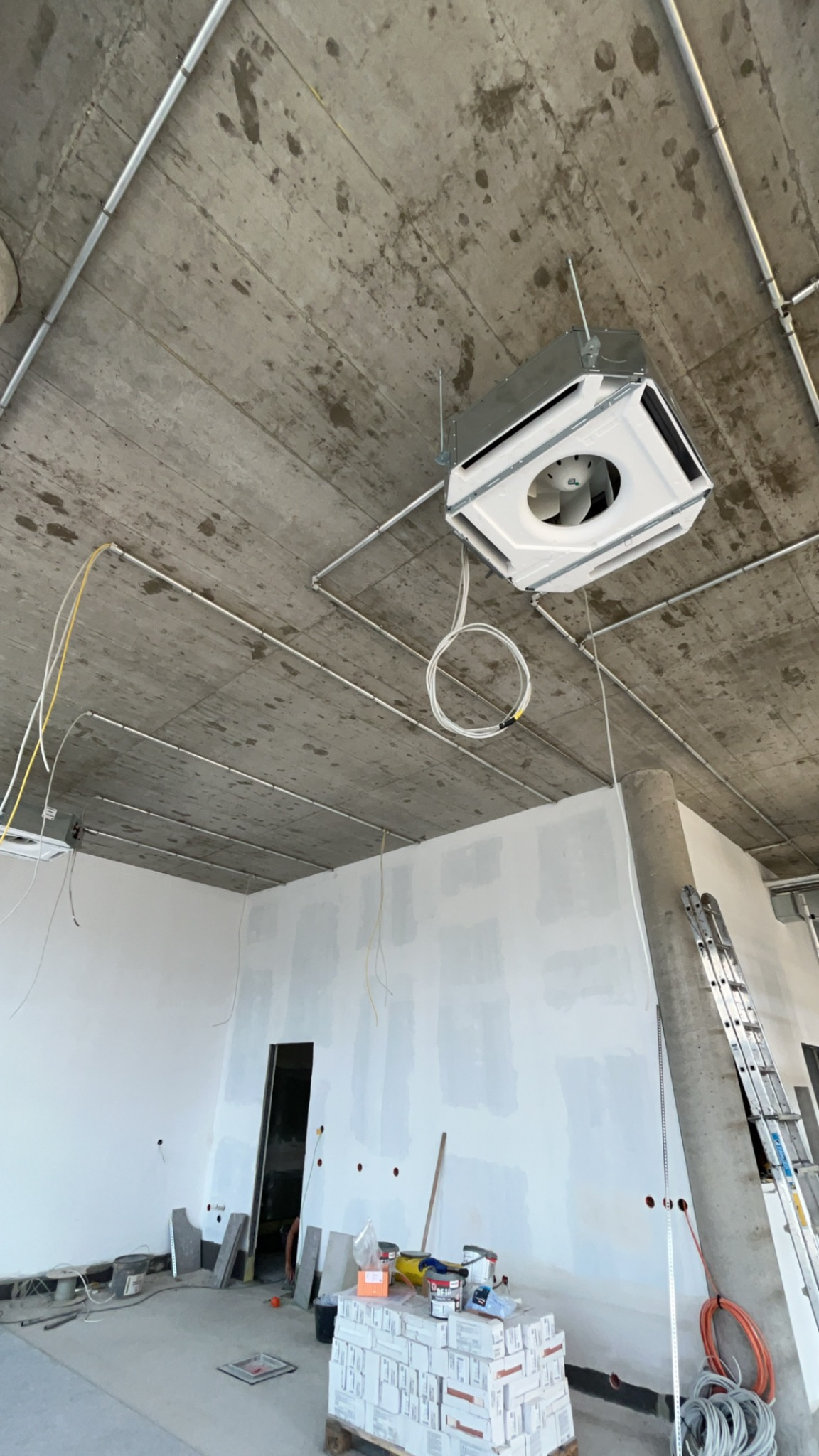 Montaż klimatyzacji kasetonowej w surowym wnętrzu. Widoczne rury instalacyjne pod sufitem i przewody elektryczne. Ściany przygotowane do malowania.