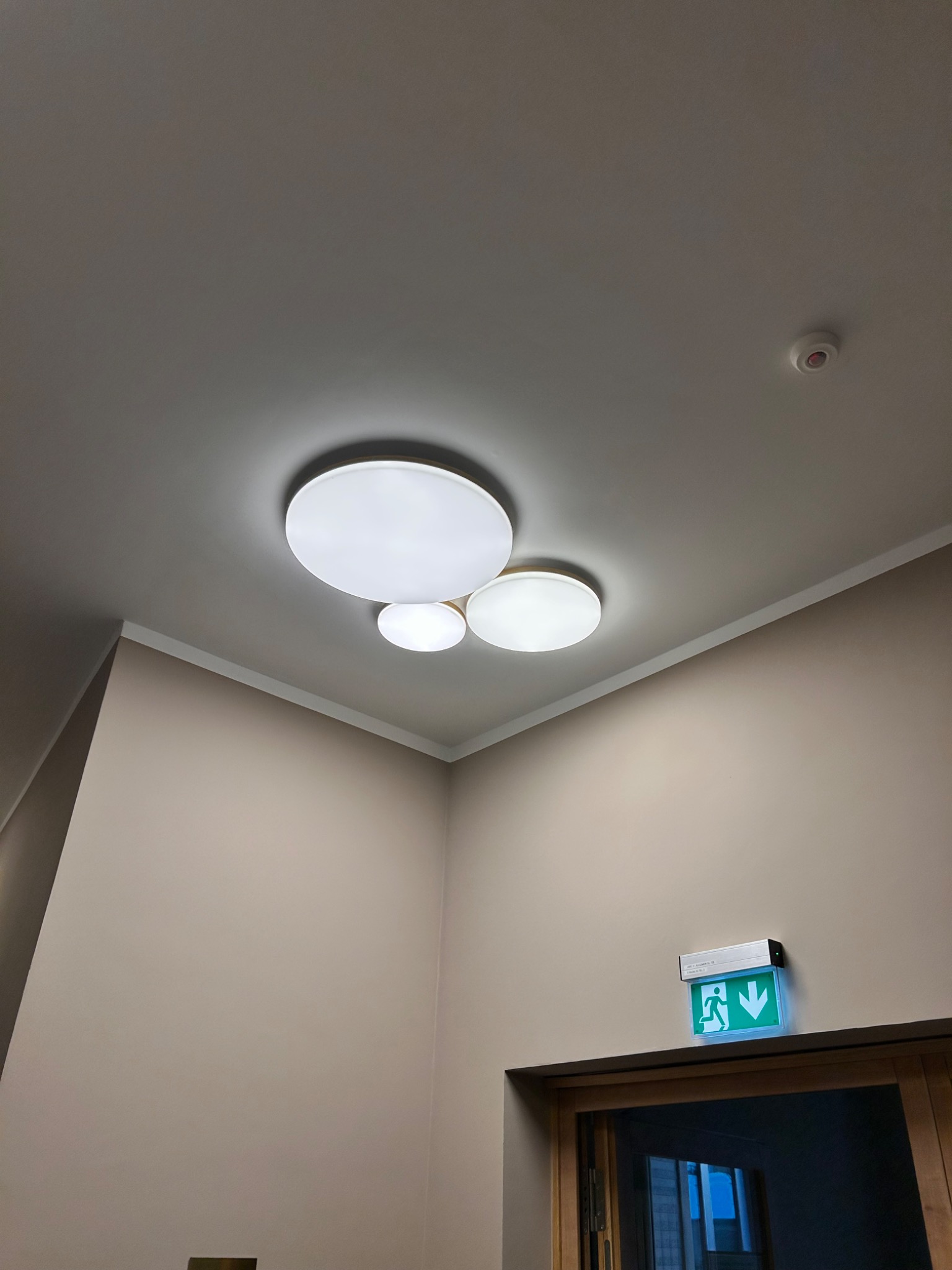 Zamontowana, nowoczesna lampa sufitowa z trzema okrągłymi kloszami, oświetlająca korytarz z beżowymi ścianami i znakiem ewakuacyjnym nad drzwiami.