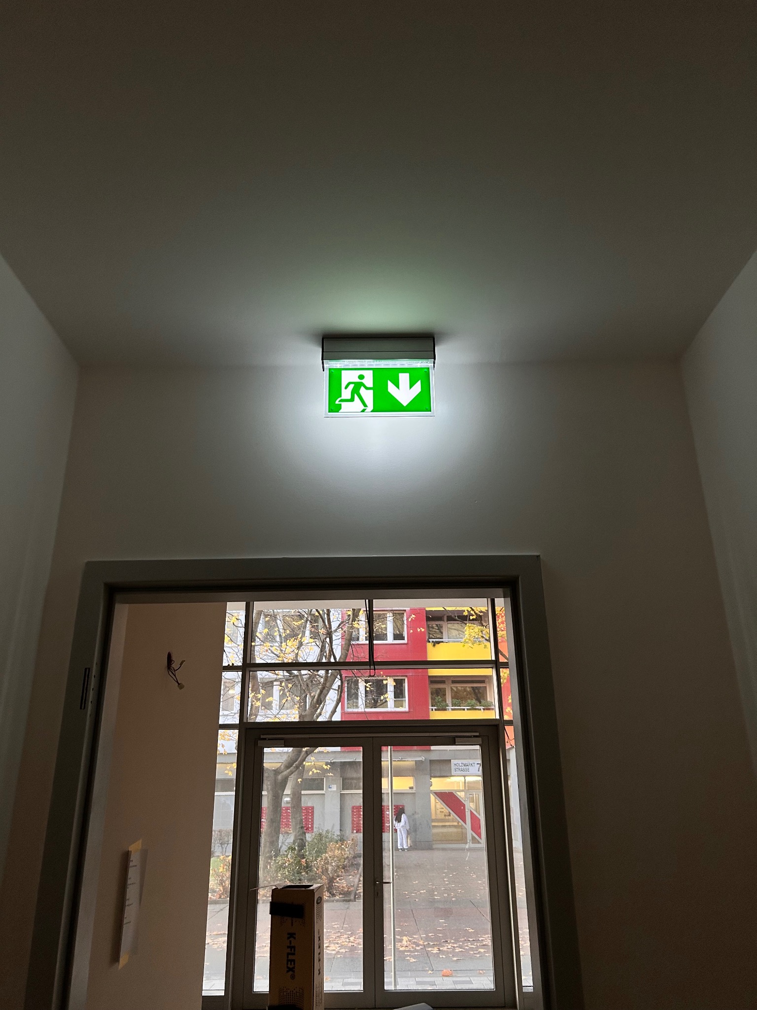 Zamontowany znak ewakuacyjny LED w Lubiszynie, widoczny nad drzwiami. Zielony piktogram osoby biegnącej i strzałka w dół. W tle budynek przez przeszklone drzwi.