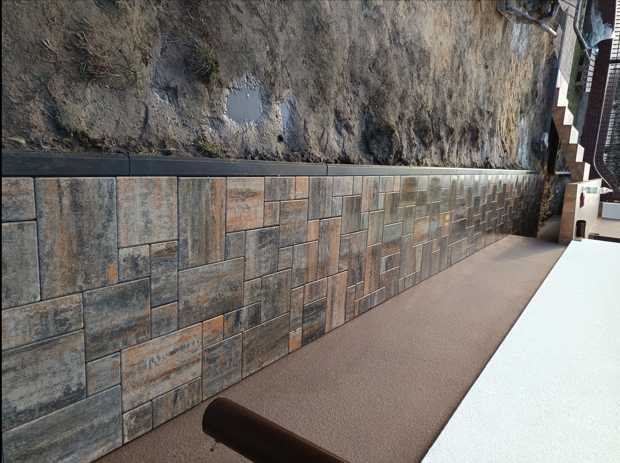 Ściana oporowa wyłożona kostką brukową w odcieniach szarości i brązu, zabezpieczająca skarpę z widoczną kałużą, z elementami schodów w tle.