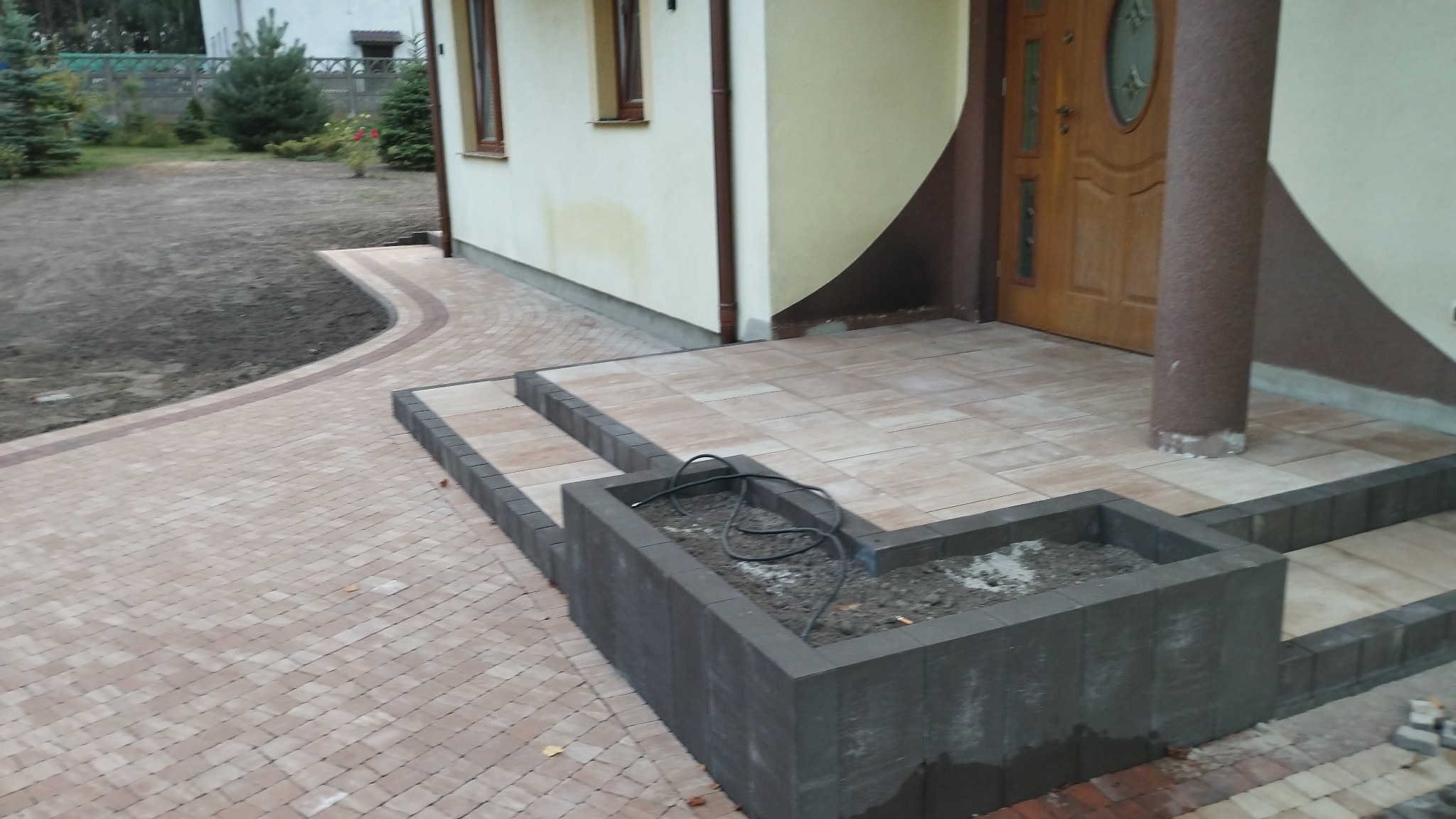 Ułożona kostka brukowa w odcieniach beżu i brązu tworząca chodnik i schody przed domem, z donicą z szarej kostki brukowej w pierwszym planie.