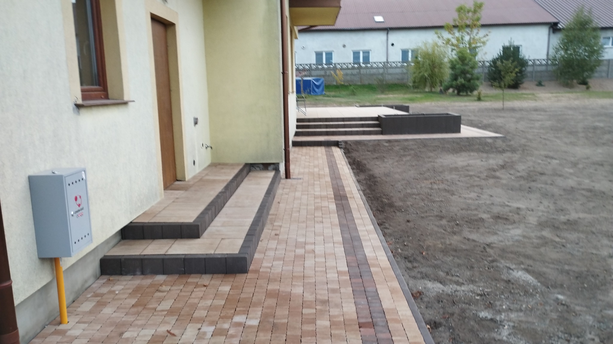 Ułożona kostka brukowa w odcieniach beżu i brązu tworząca ścieżkę i schody przed domem z szarą skrzynką gazową na żółtej rurze