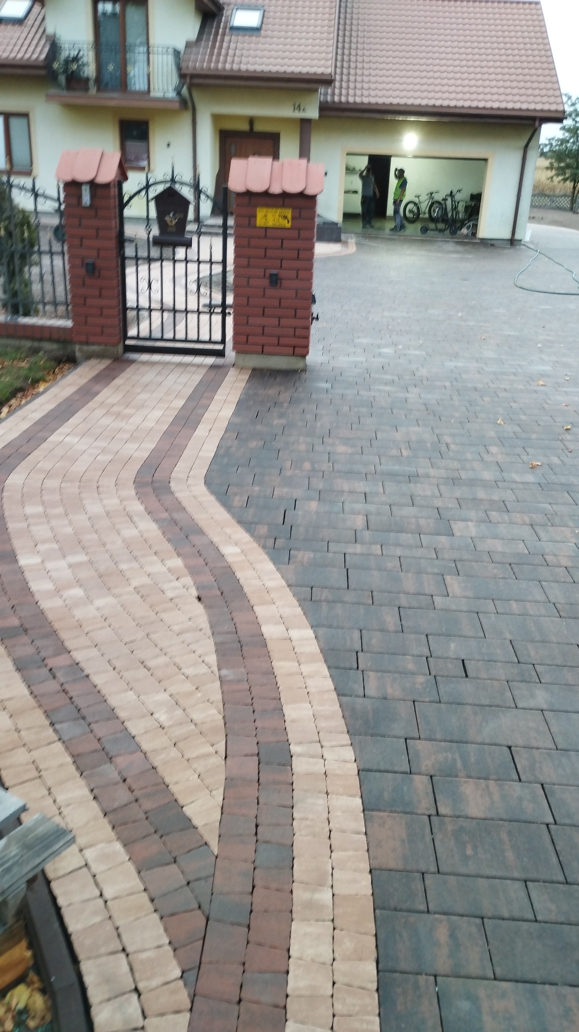 Ułożona z dwóch rodzajów kostki brukowej ścieżka w kształcie fali prowadząca od furtki do garażu, z widocznymi fragmentami domu w tle.