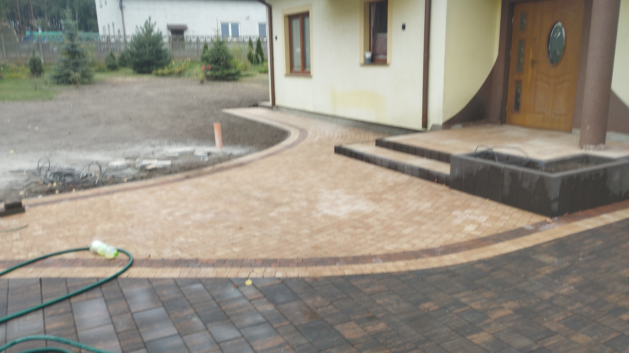 Ułożona beżowo-brązowa kostka brukowa tworząca ścieżkę i podjazd przed domem z drewnianymi drzwiami wejściowymi, widoczny fragment trawnika z młodymi drzewkami i zielony wąż ogrodowy.