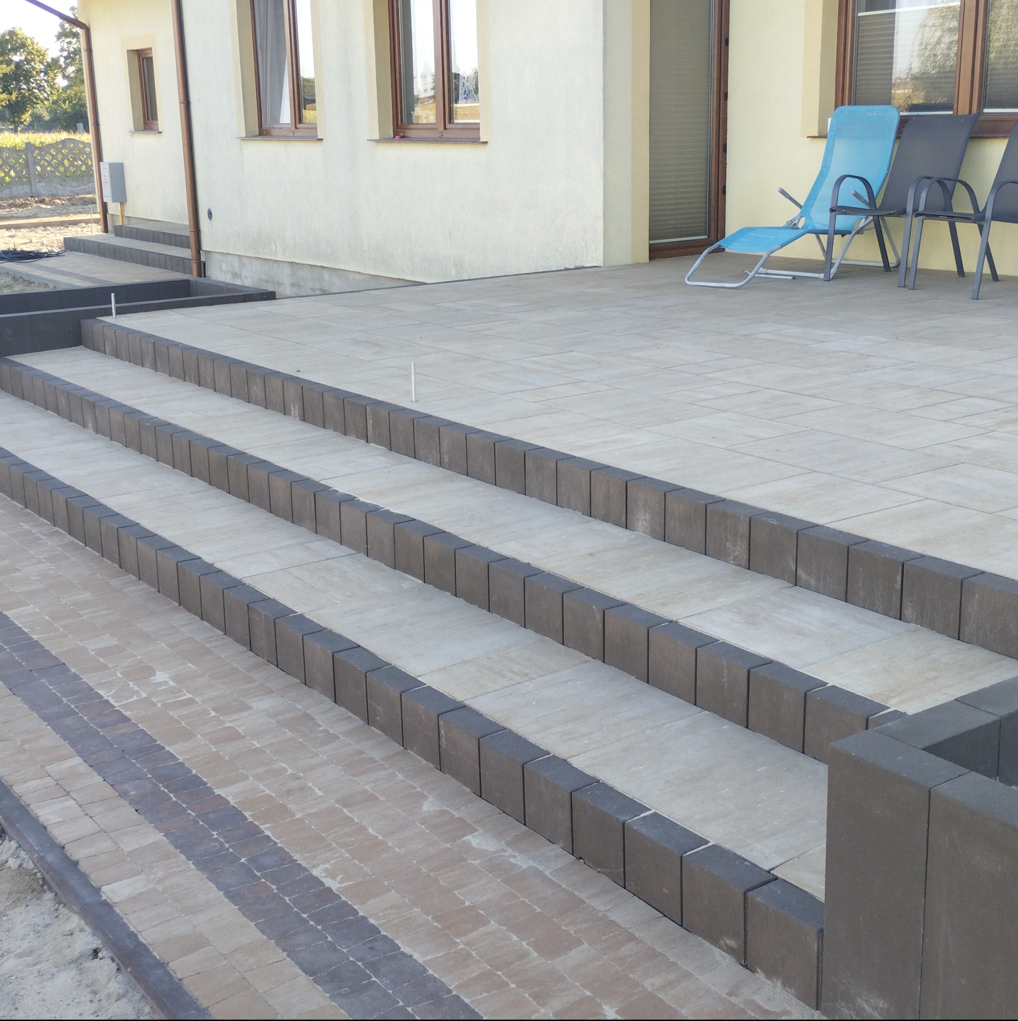 Schody z kostki brukowej prowadzące do domu, z widoczną częścią tarasu i meblami ogrodowymi w tle.