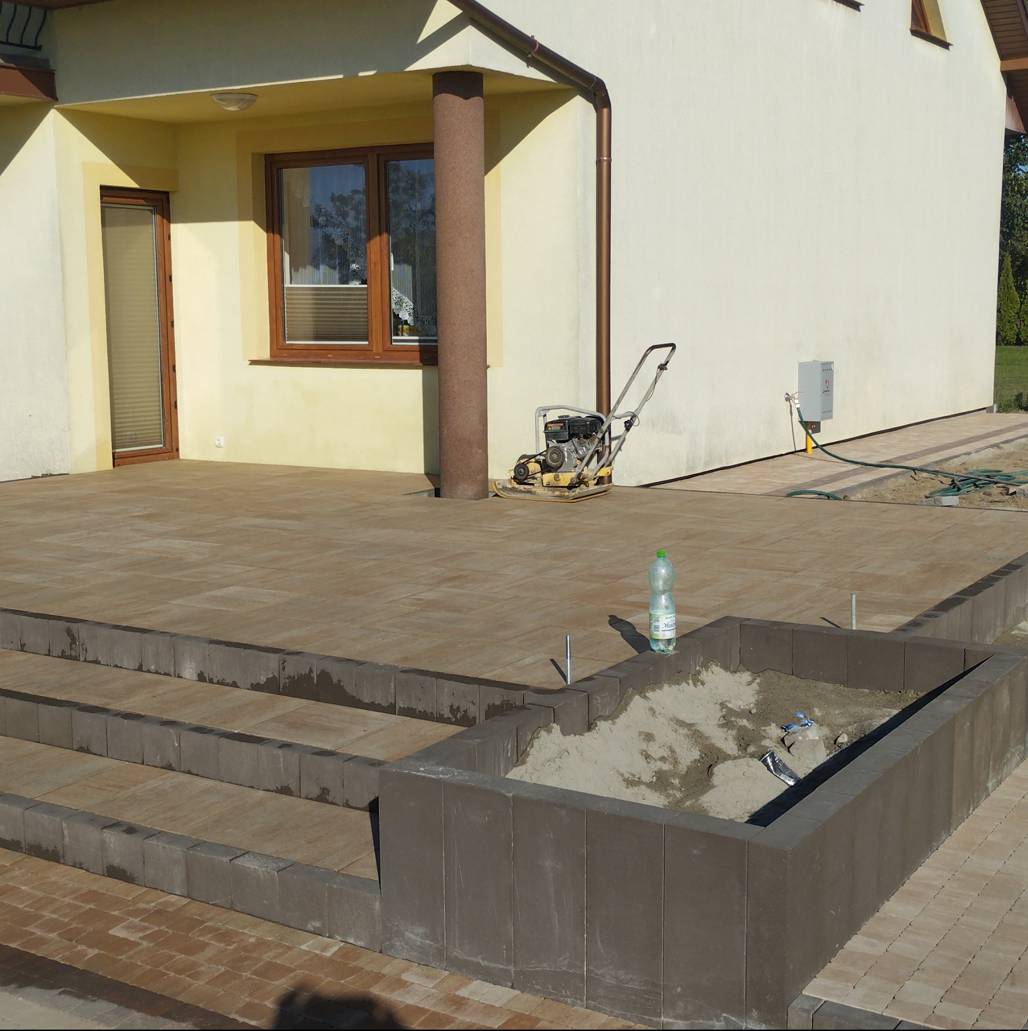 Ułożona kostka brukowa na tarasie z schodami i obrzeżem rabaty kwiatowej, ubijarka wibracyjna w tle, butelka wody mineralnej na kostce, częściowo wypełniona ziemią rabata.