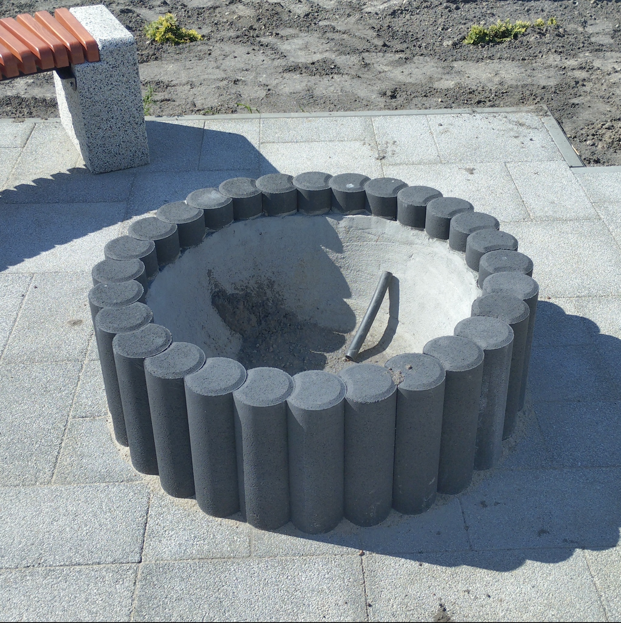 Okrągła rabata z betonowych elementów na tle kostki brukowej i ziemi, widoczny fragment ławki.