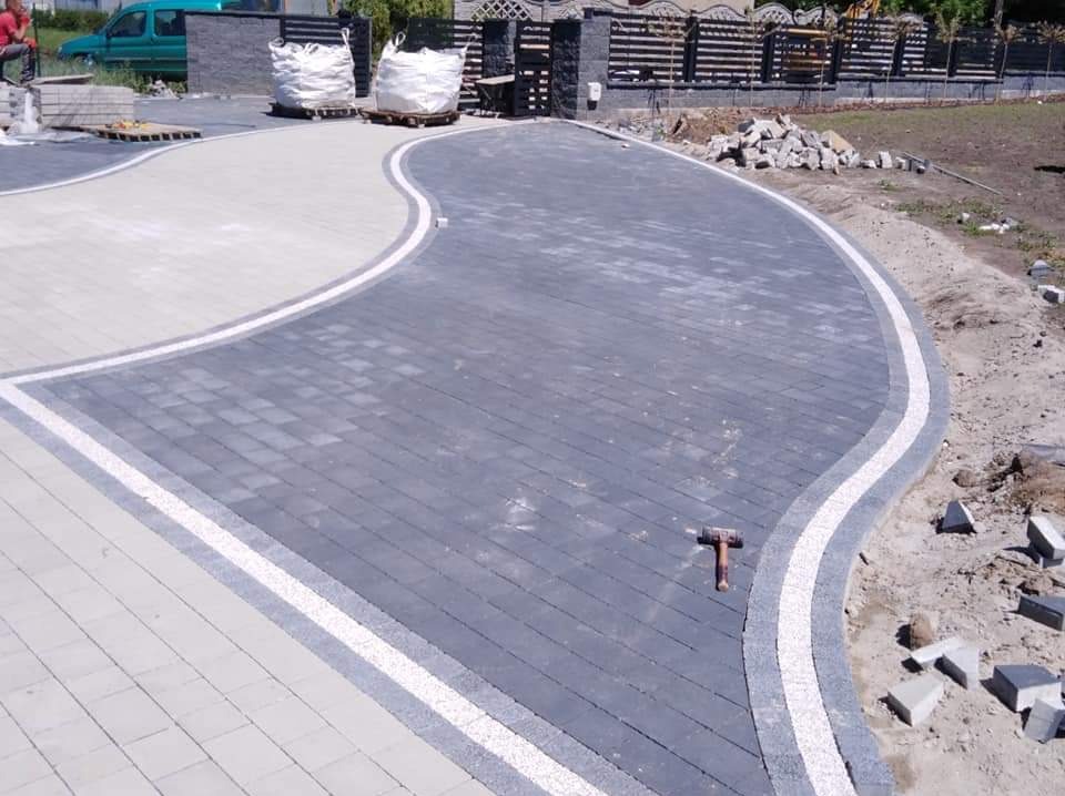 Układanie kostki brukowej w kształcie litery S z ciemnoszarej kostki obramowanej jasnoszarą kostką, widoczny młotek brukarski, worki z materiałem i częściowo ogrodzenie.