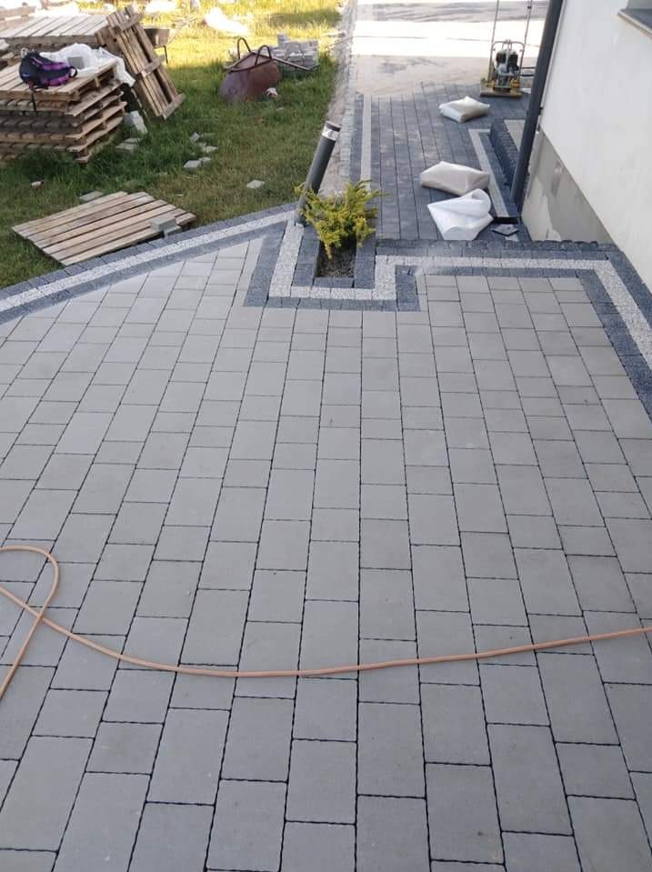 Ułożona szara kostka brukowa z ciemniejszą obwódką i miejscem na roślinę, widoczne palety i narzędzia w tle.