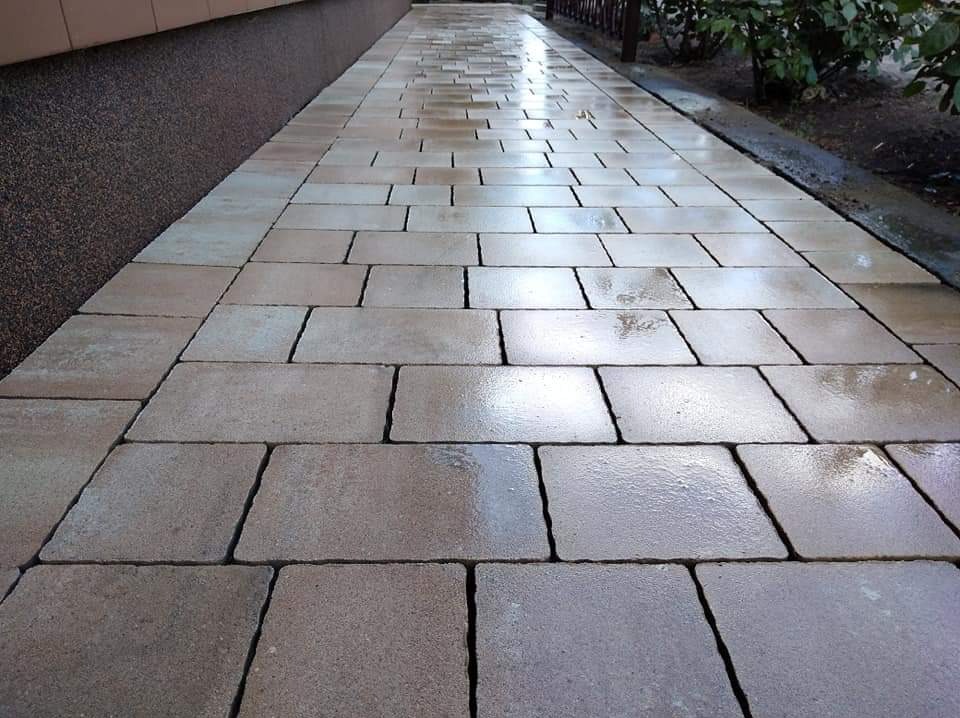 Świeżo ułożony chodnik z prostokątnej, beżowej kostki brukowej, mokry od deszczu, z widocznym fragmentem elewacji budynku i roślinności w tle.