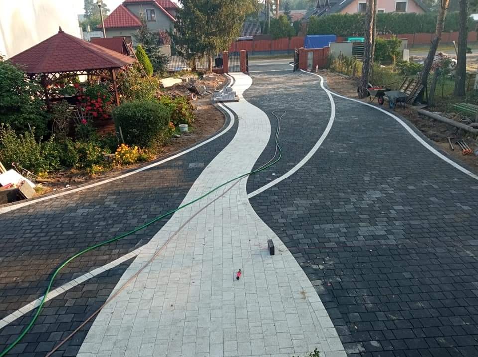 Szeroki, kręty podjazd z kostki brukowej w dwóch kolorach, z białymi obrzeżami, prowadzący do bramy wjazdowej, otoczony zielenią i elementami architektury ogrodowej.