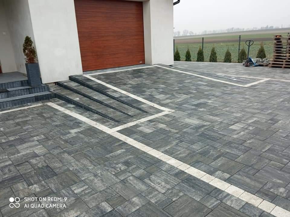 Ułożona szara kostka brukowa z białymi akcentami na podjeździe w Śleszynie, widoczne schody wejściowe do domu i brama garażowa.