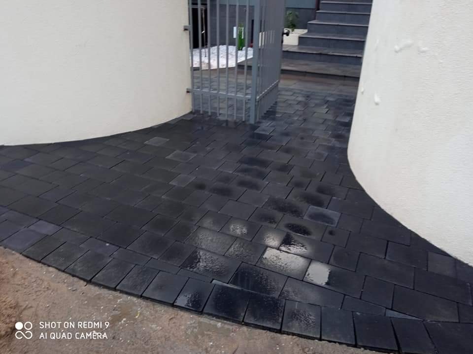 Świeżo ułożona, czarna kostka brukowa tworząca zakrzywiony chodnik prowadzący do metalowej bramki i schodów; widoczne mokre plamy na powierzchni kostki.