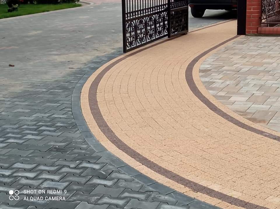 Ułożona kostka brukowa w dwóch odcieniach szarości i beżu, tworząca łukowaty podjazd z dekoracyjnym obramowaniem, prowadzący do bramy wjazdowej.