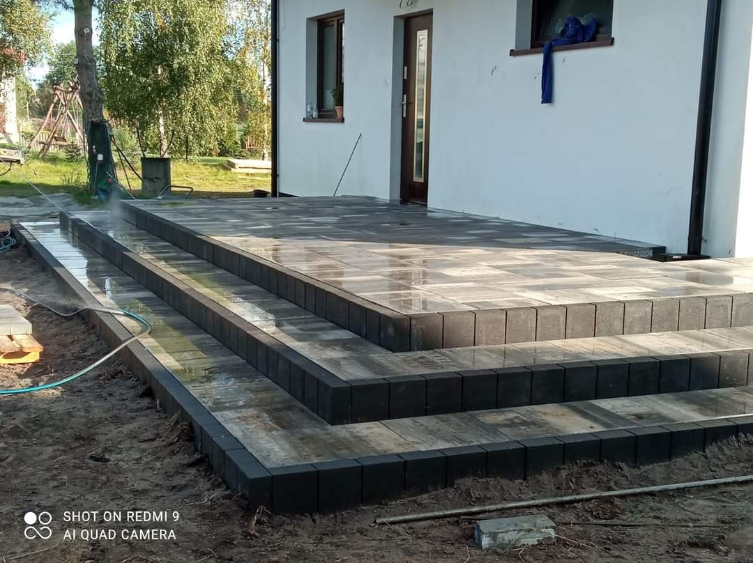 Schody i taras przed domem wyłożone szarą kostką brukową, czyszczone wodą pod ciśnieniem, widok na elewację budynku.