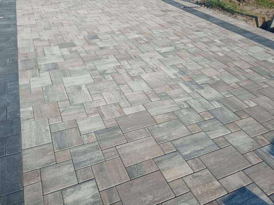 Ułożona nawierzchnia z szarej, prostokątnej kostki brukowej o zróżnicowanych odcieniach, obrzeża z ciemniejszej kostki, widoczne fragmenty trawy w tle.