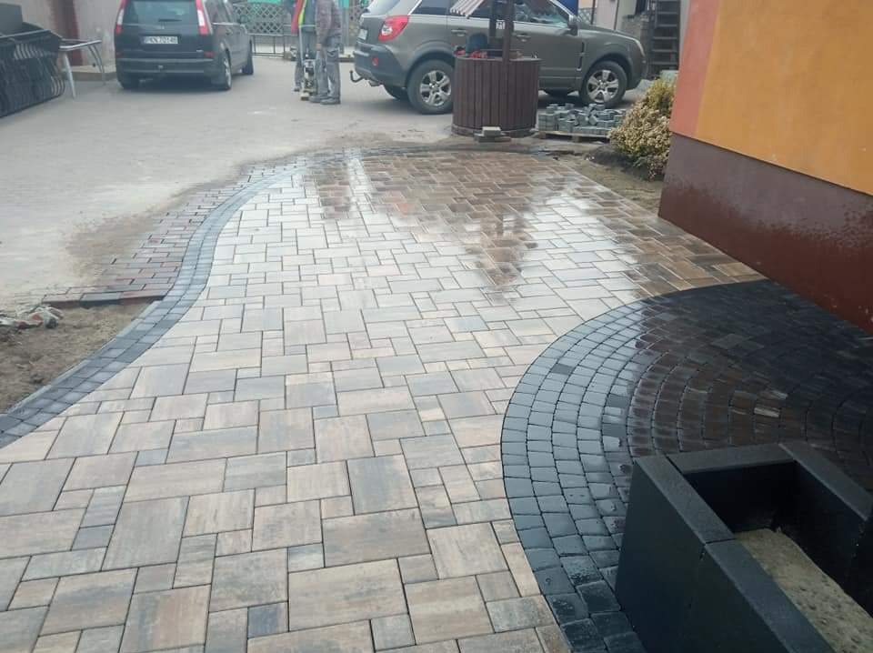 Świeżo ułożona nawierzchnia z kostki brukowej w odcieniach szarości i beżu, z dekoracyjnym obramowaniem z ciemnej kostki, odbijająca światło po deszczu, z widocznymi dwoma samochodami w tle.