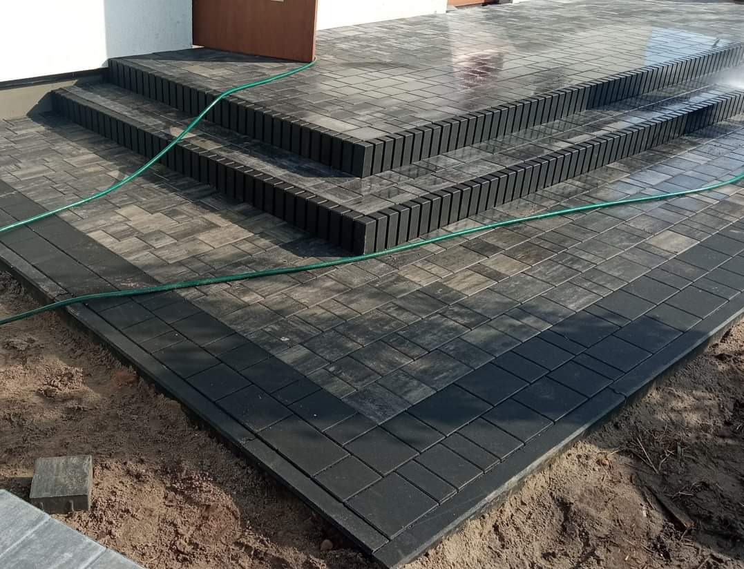 Ułożona, szara kostka brukowa tworząca schody wejściowe i chodnik, obramowana ciemniejszymi kostkami, z zielonym wężem ogrodowym leżącym na powierzchni.