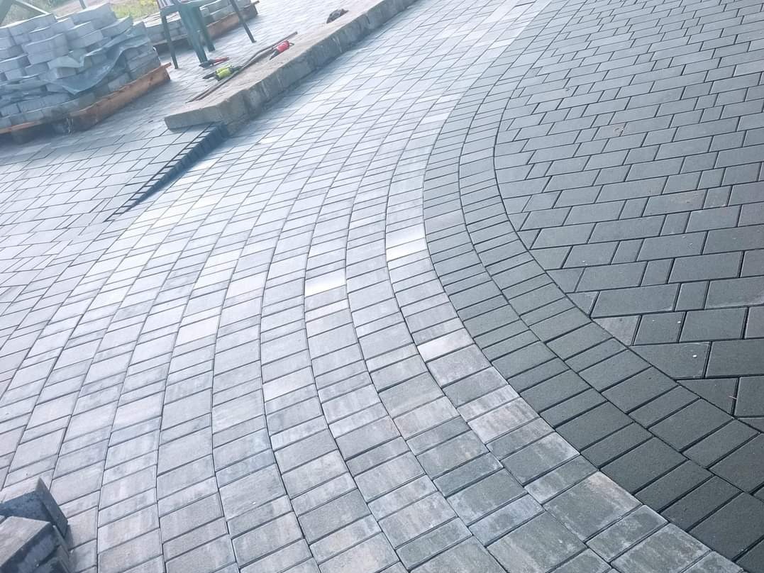 Ułożona nawierzchnia z kostki brukowej w dwóch odcieniach szarości, tworząca łukowaty wzór. Częściowo widoczne narzędzia i paleta z kostką w tle.