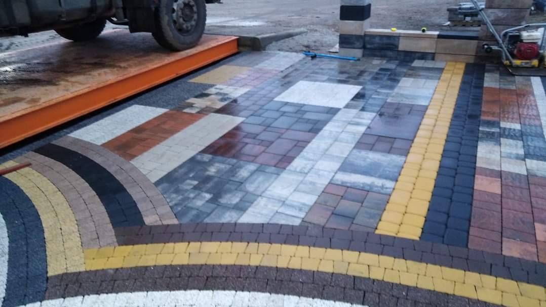 Układanie wielokolorowej kostki brukowej z wzorem łukowym, widoczna ubijarka wibracyjna i fragment ciężarówki na rampie.