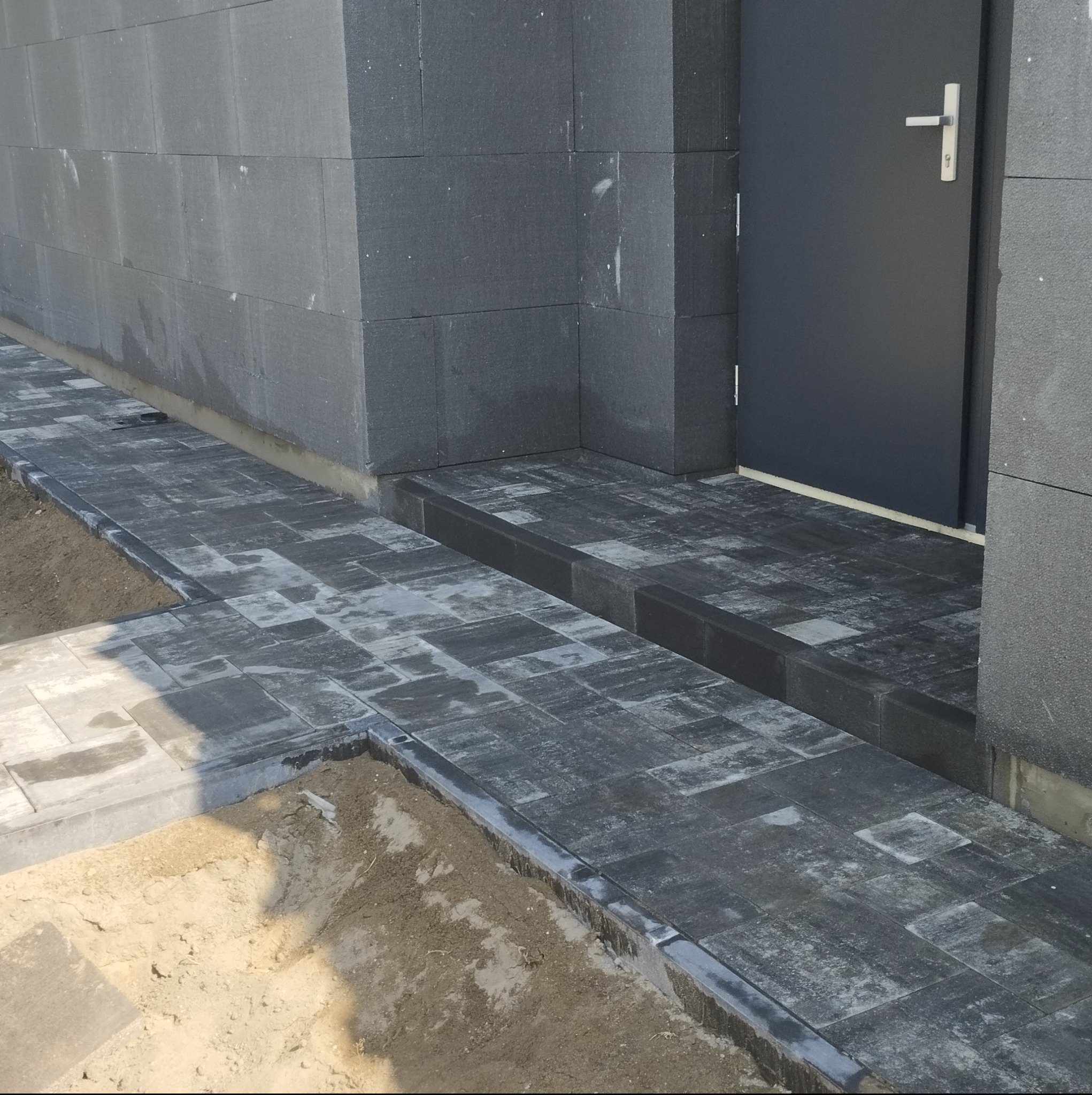Nowa, ciemnoszara nawierzchnia z kostki brukowej, kontrastująca z surową, szarą ścianą budynku i ciemnymi drzwiami wejściowymi; widoczny fragment przygotowanego podłoża z piaskiem.