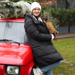 Olena Dankiv - Uśmiechnięta kobieta w zimowej kurtce i czapce, oparta o czerwony samochód, trzyma bukiet z gałązek iglastych i czerwonych jagód, na dachu auta choinka w siatce.