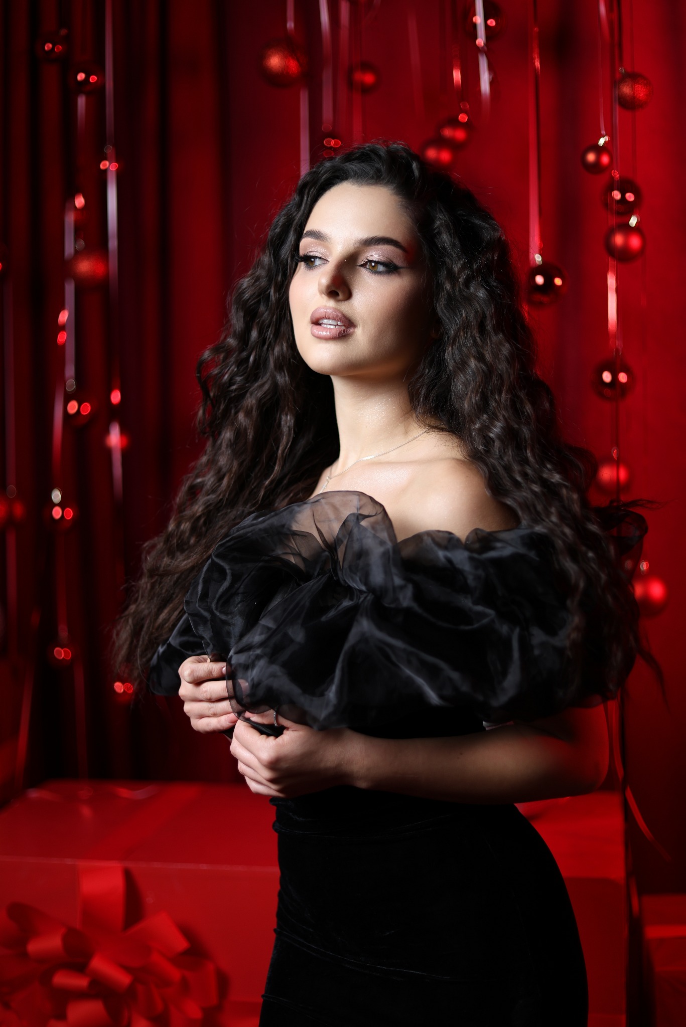 Portret kobiety w czarnej sukni z bufiastymi rękawami na tle czerwonej dekoracji świątecznej z bombkami i prezentami.