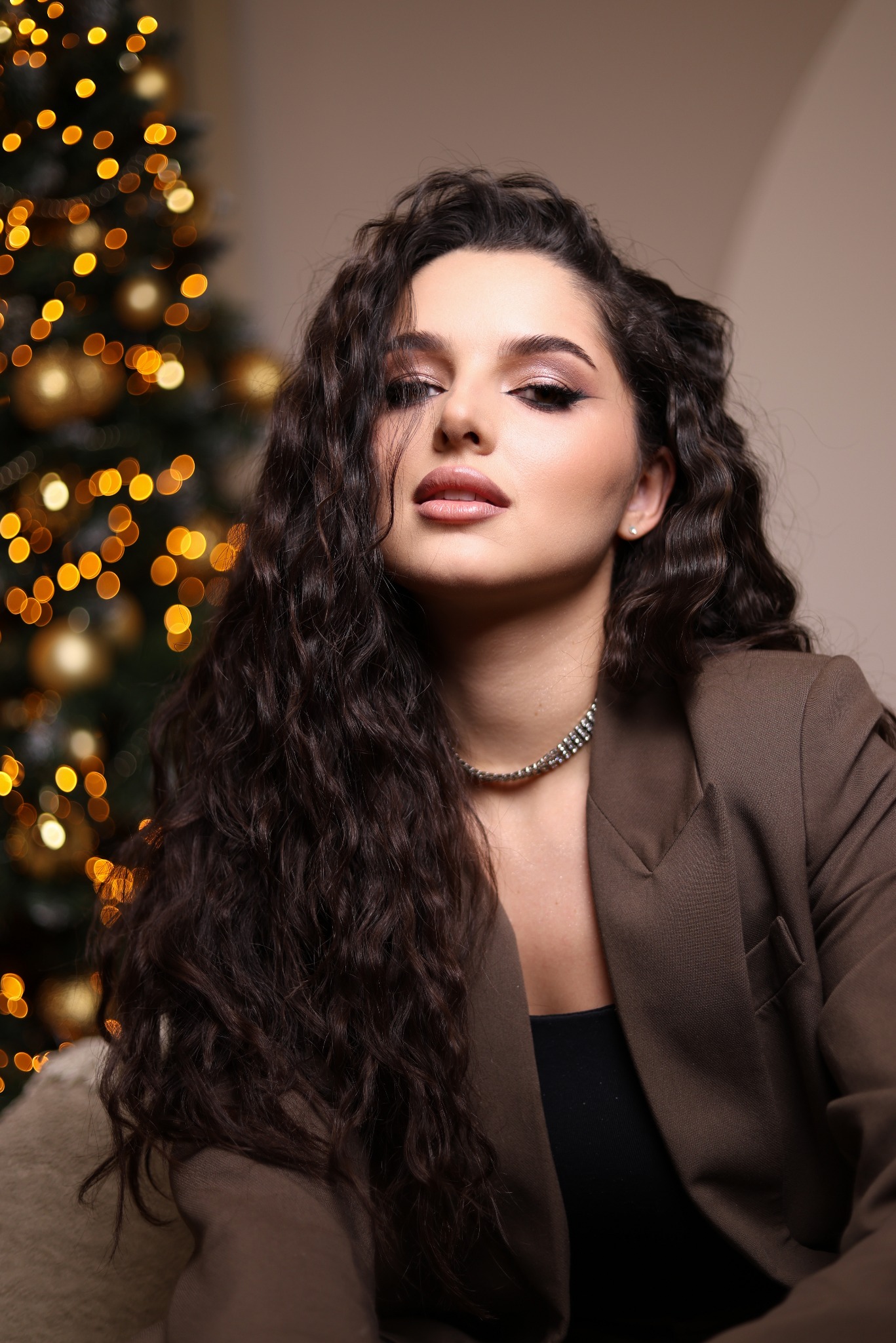 Portret kobiety w brązowej marynarce, z długimi, ciemnymi, kręconymi włosami i wyrazistym makijażem, pozującej na tle choinki z bokeh.