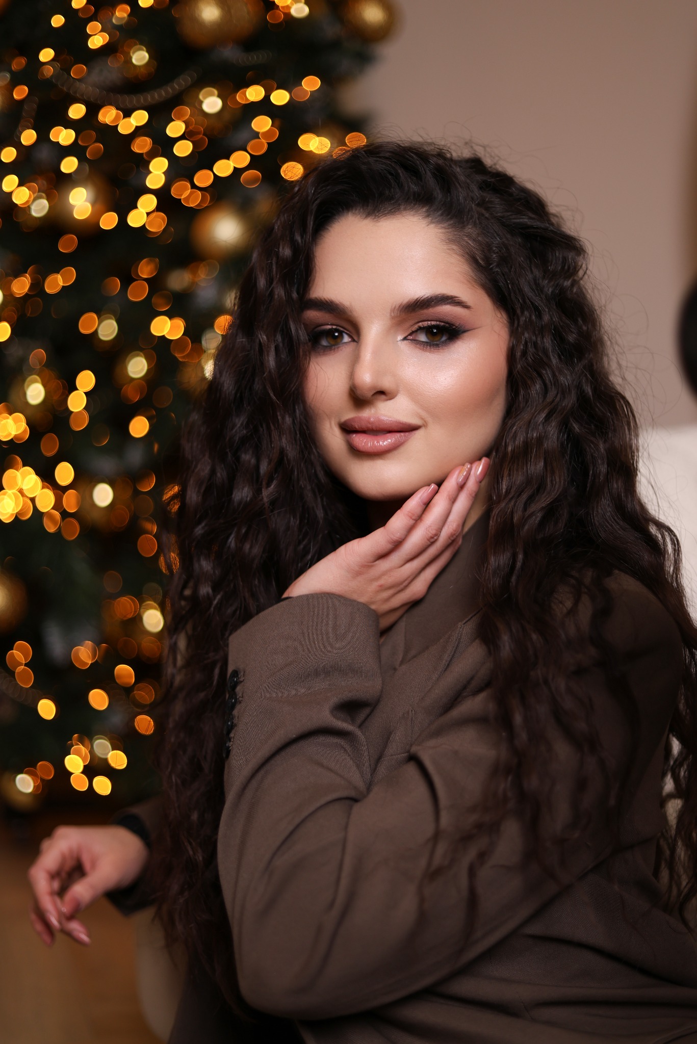 Portret kobiety z długimi kręconymi włosami w brązowej marynarce, pozującej na tle rozświetlonej choinki.