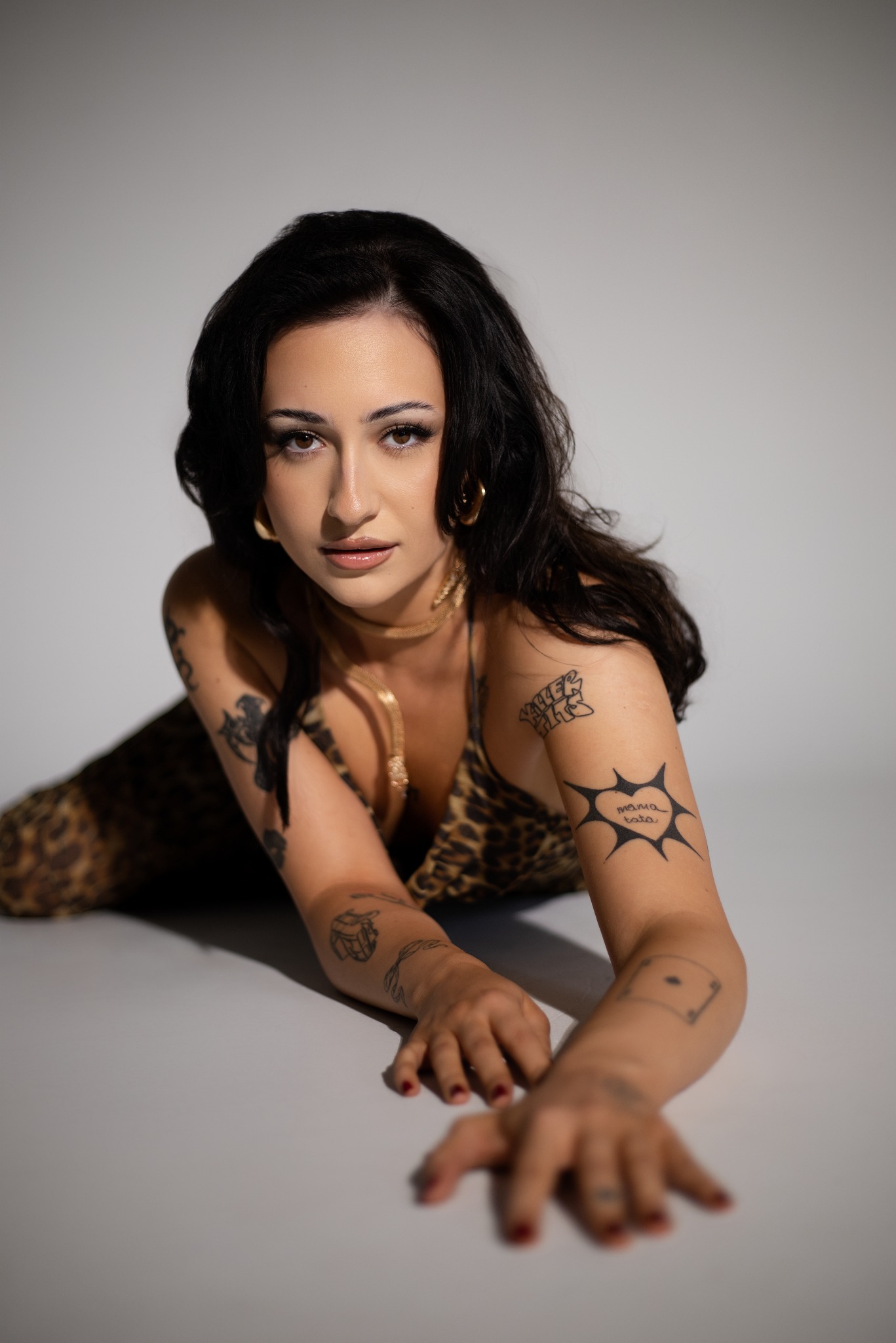 Portret kobiety w sukience w panterkę, leżącej na podłodze, z widocznymi tatuażami na ramionach i złotą biżuterią, na jasnoszarym tle.