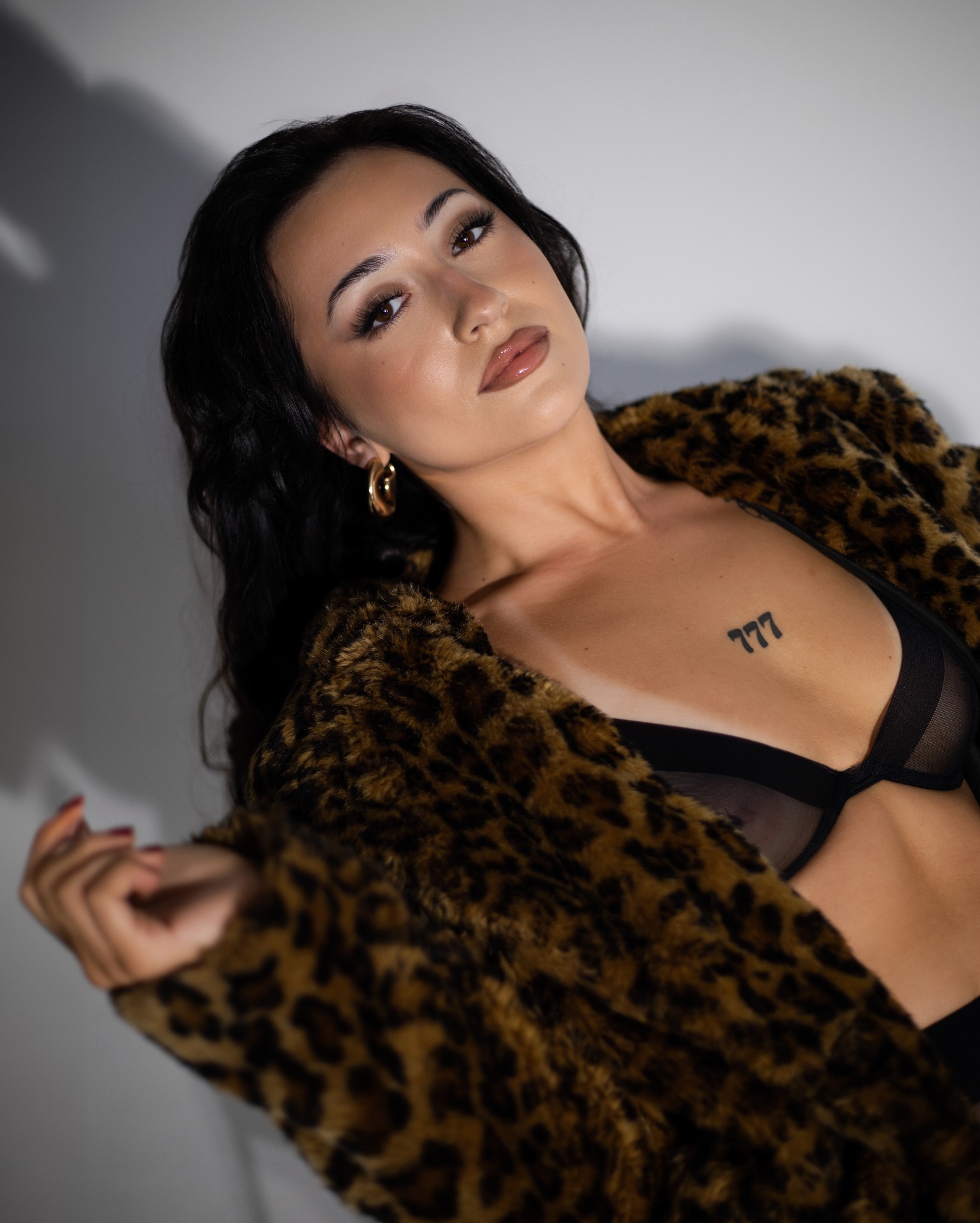 Portret kobiety w futrze w panterkę, z widocznym tatuażem '777' na dekolcie i złotymi kolczykami w kształcie koła, pozującej na tle jasnej ściany z cieniem.